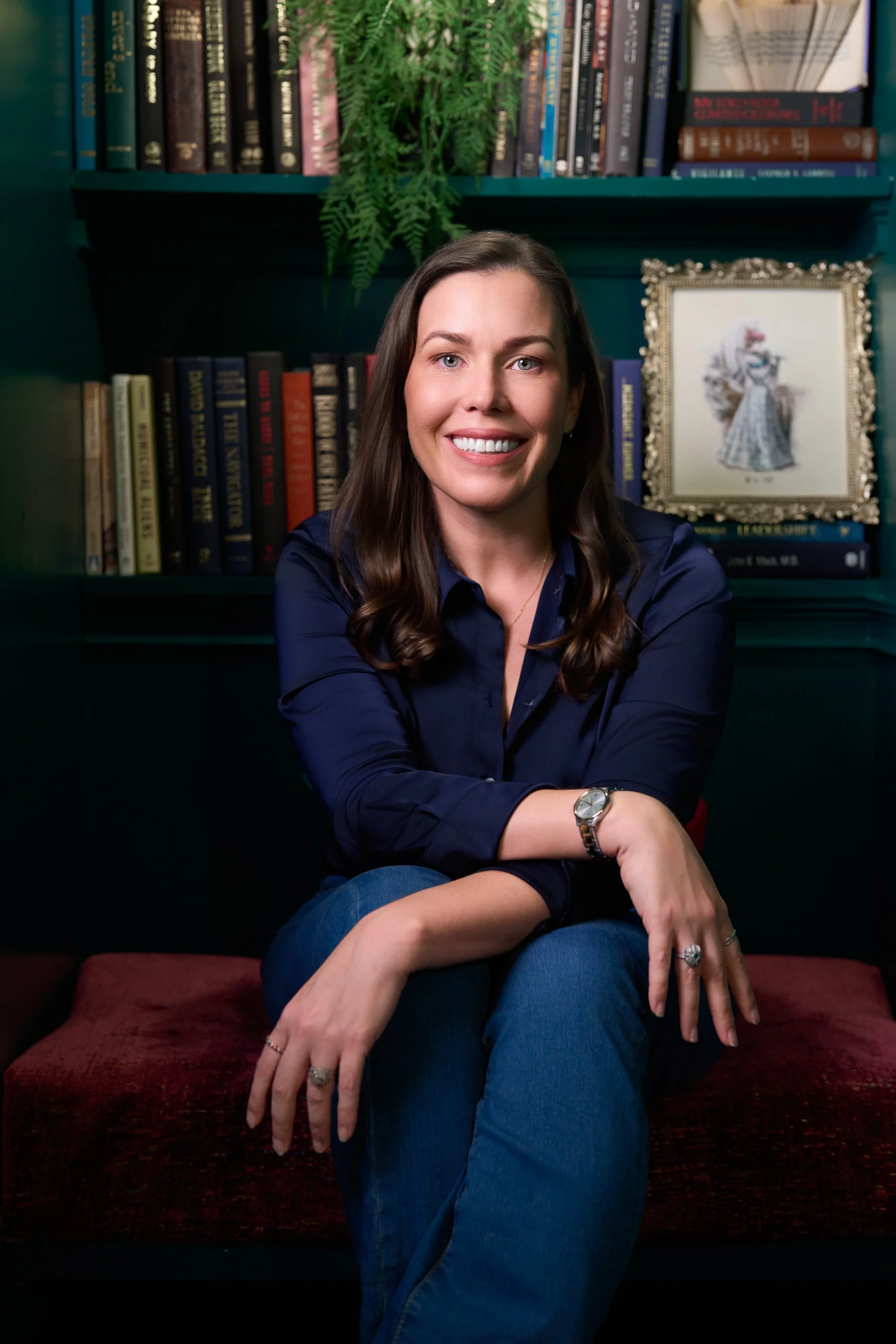 Amy Cherven with long brown hair and a bright smile sitting on a red velvet couch in front of a blue bookshelf filled with books, a framed picture of a woman in a dress, and a green fern plant.