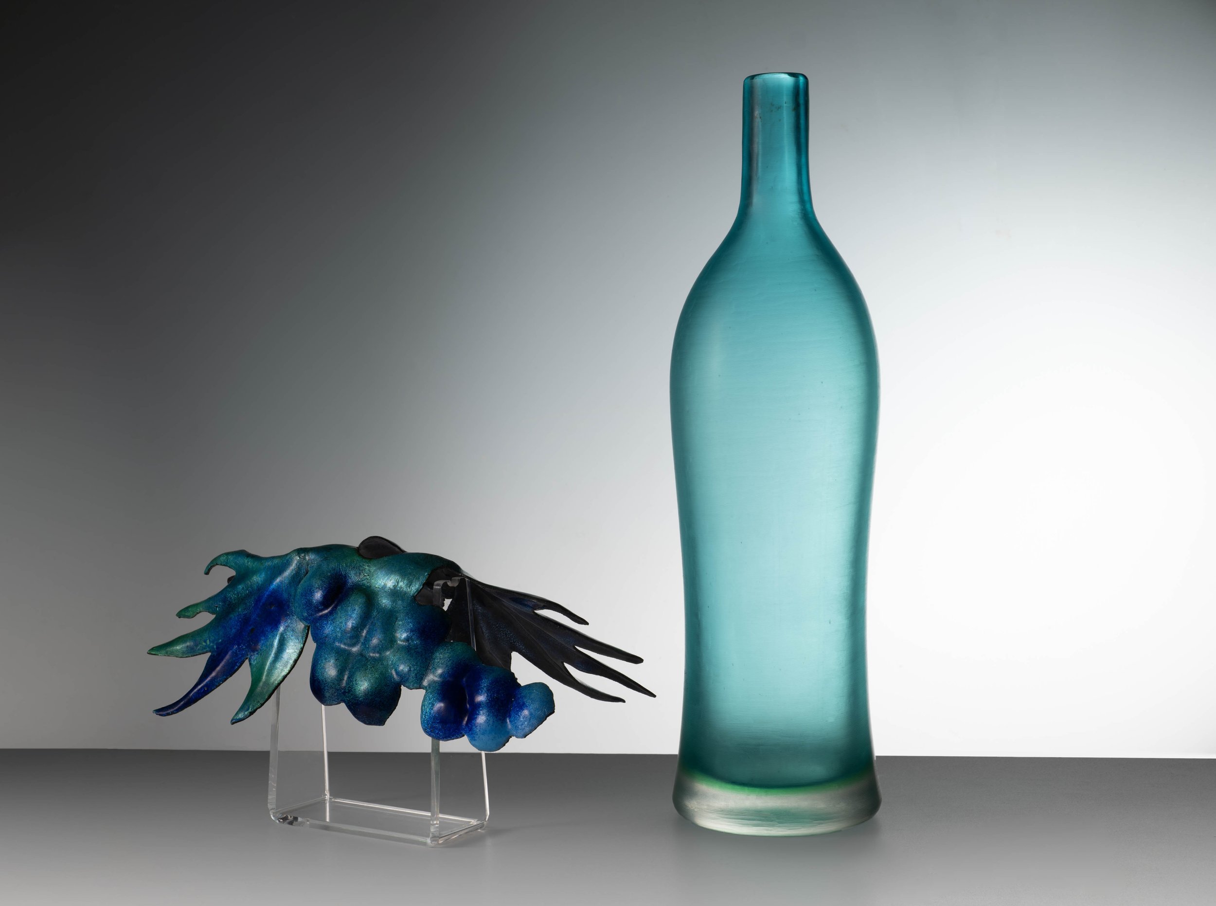 Un vaso di vetro blu accanto a una scultura astratta in vetro che ricorda un'ala o una foglia con tonalità blu e verdi, esposta su uno sfondo neutro. Museo del Vino Lungarotti 