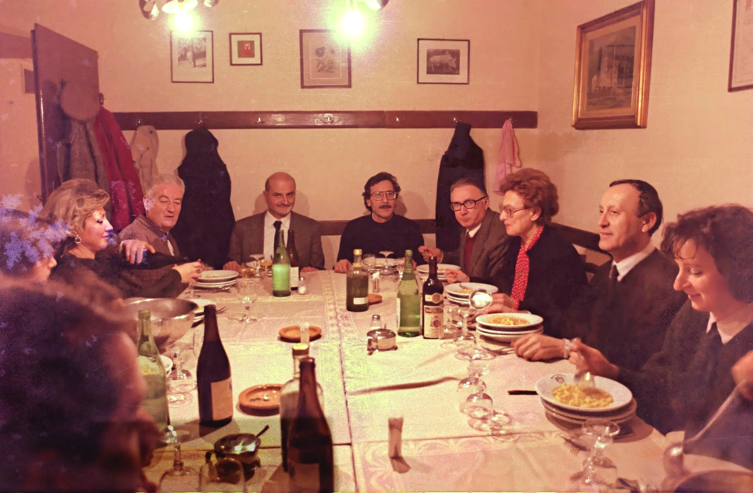 People sitting around a long dining table in a cozy restaurant or home with framed pictures on the wall and hanging lights. Food, drinks, and tableware are on the table.