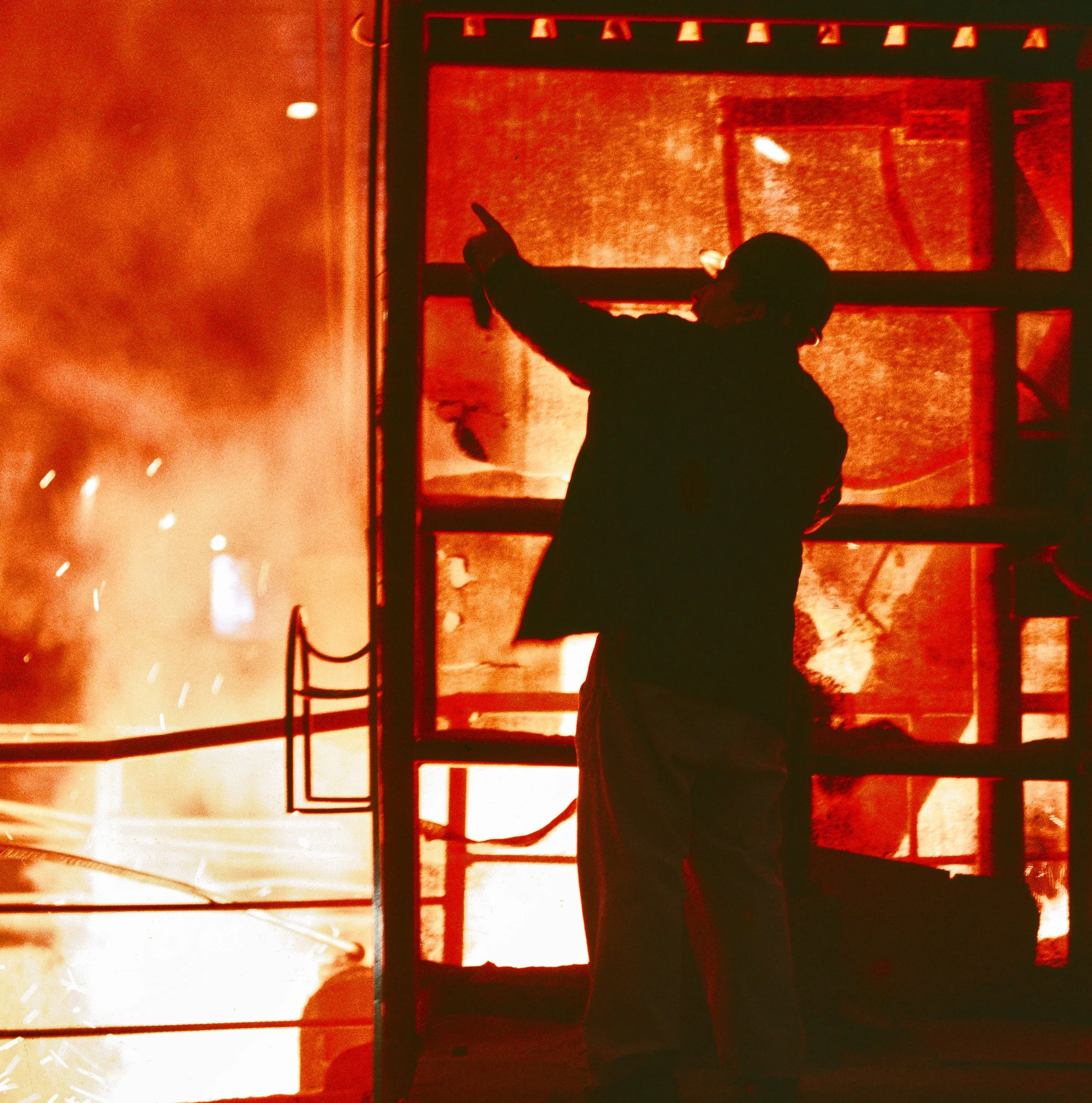 A person silhouette pointing at a screen displaying a red and black graph or chart, with the scene filled with orange and red hues and sparks or debris in the background.