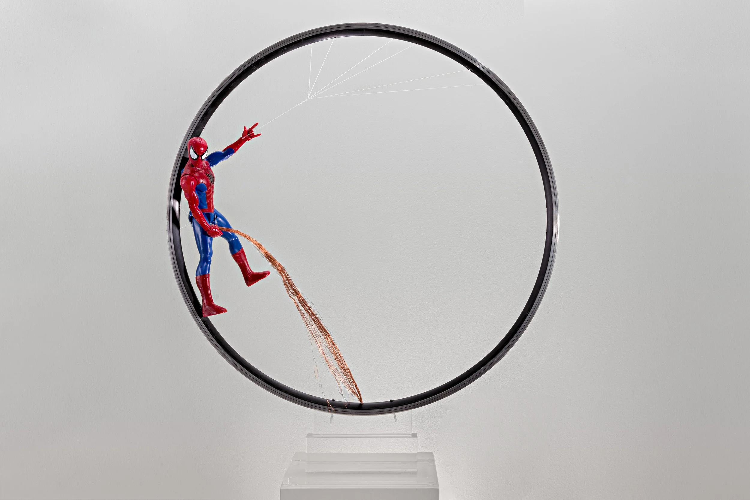 A Spiderman action figure hanging from thin Scultura di fili all'interno di una cornice circolare nera, con una scia di filo rosso sotto di lui, esposti su un supporto trasparente su uno sfondo bianco uniforme Spider Man appeso. 