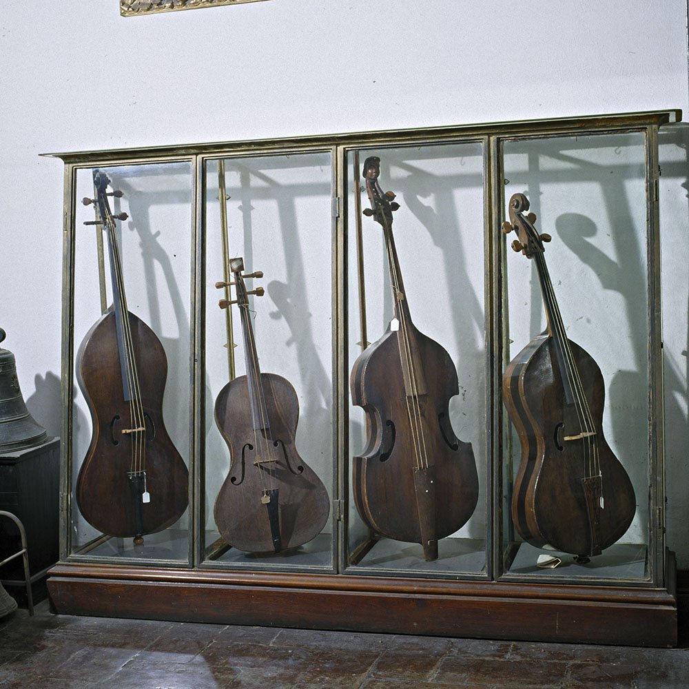 Museo civico di Modena. Quattro strumenti a corda d'epoca, tra cui violoncelli e contrabbassi, esposti in una teca di vetro su una parete bianca.