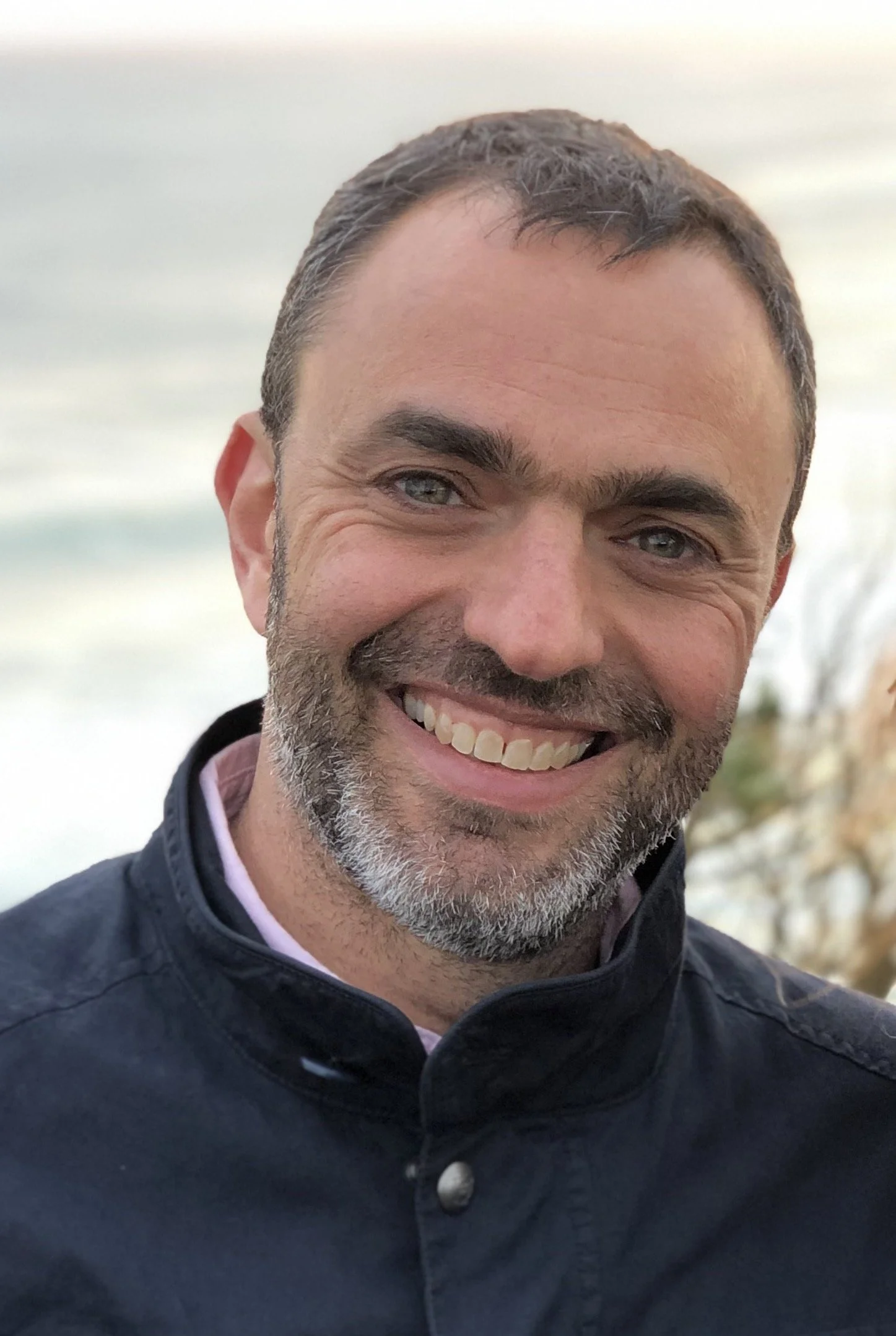 Close-up photo of a smiling man with short dark hair, a beard, and light-colored eyes, wearing a dark jacket with a high collar.