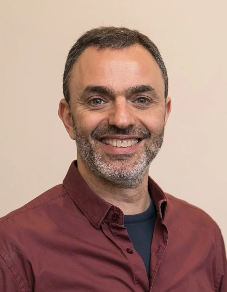 A smiling man with short dark hair, beard, wearing a maroon button-up shirt and a dark T-shirt against a plain beige background.