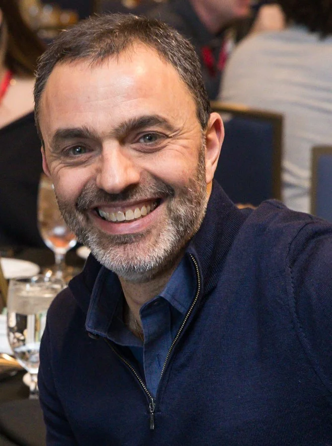 Close-up of a smiling man with short dark hair and a beard, wearing a navy blue jacket over a blue shirt, sitting at a table in a restaurant or event setting.