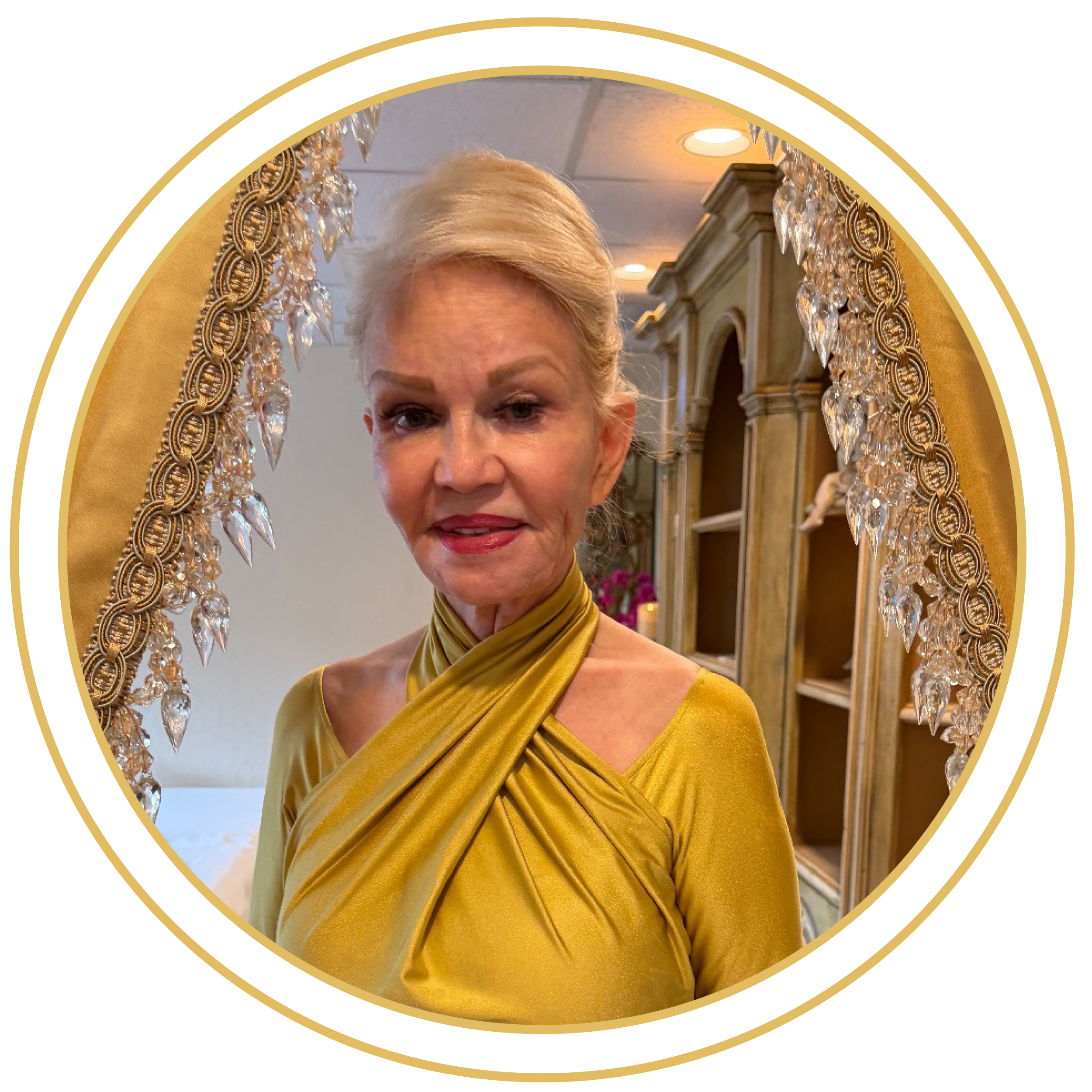 A woman with blonde hair and red lipstick wearing a gold dress with a pleated fabric pattern, standing in front of a decorative mirror with crystal embellishments, in a warmly lit room.