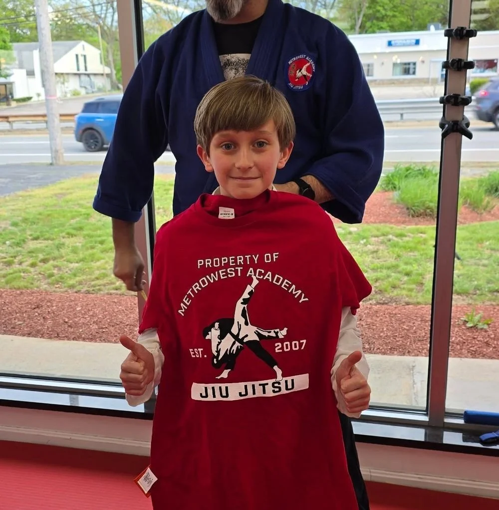 A student holding up a Property of Metrowest Academy t-shirt with Sensei Paul behind him