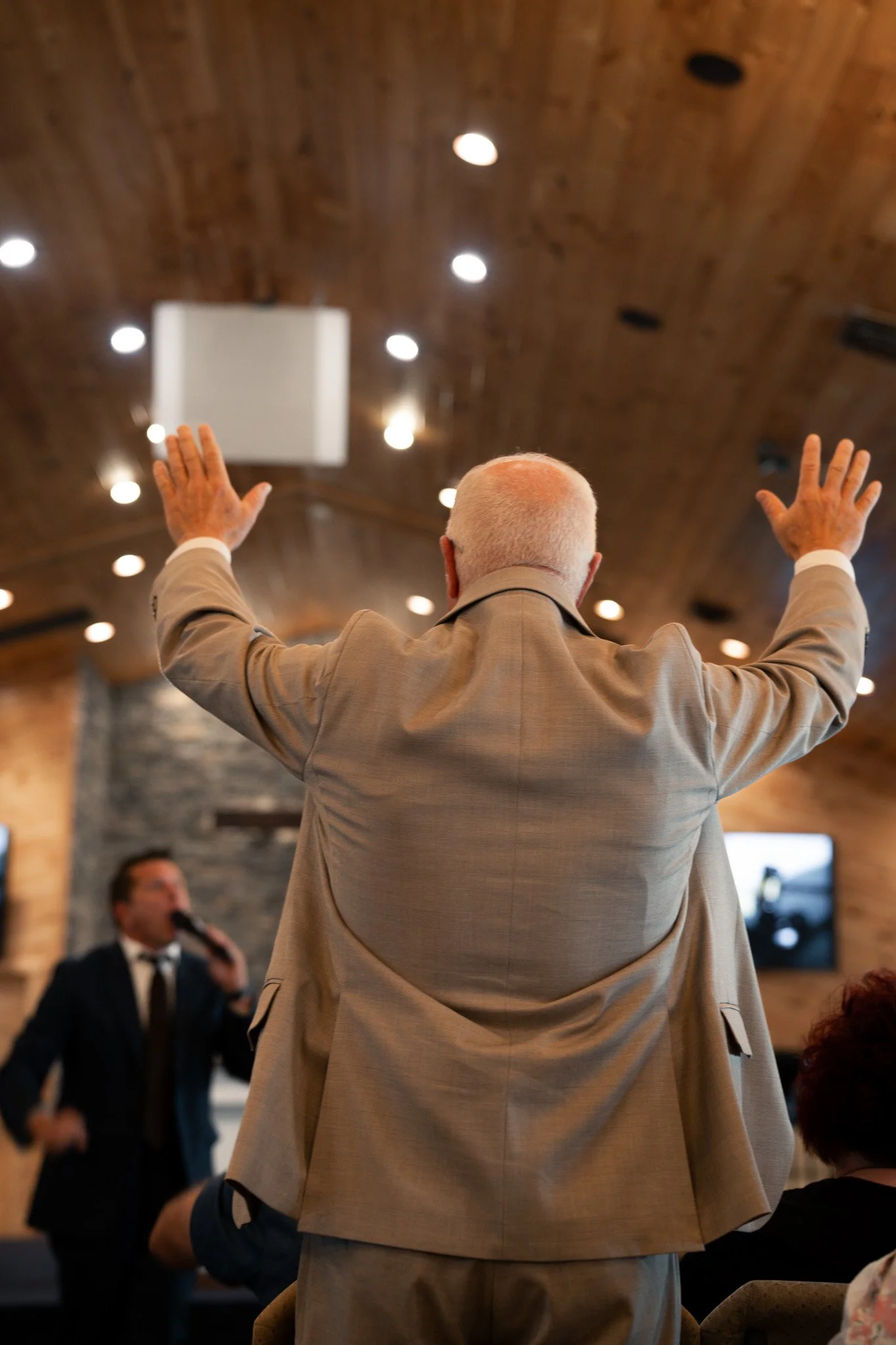 An elderly man with white hair and dressed in a beige suit is standing with his back to the camera, raising his hands in the air during a speech or presentation in a wooden-paneled room with ceiling lights. A man in a black suit and bow tie is speaki