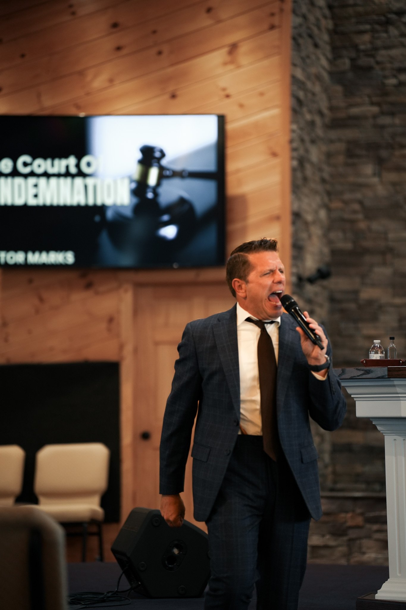 A man in a suit passionately singing into a microphone during a speech or performance, with a wooden wall and a large screen displaying text behind him.