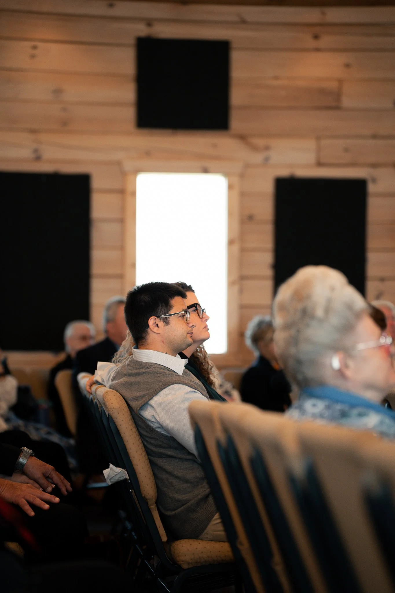 People sitting in chairs attending a conference or seminar in a room with wooden walls and windows.