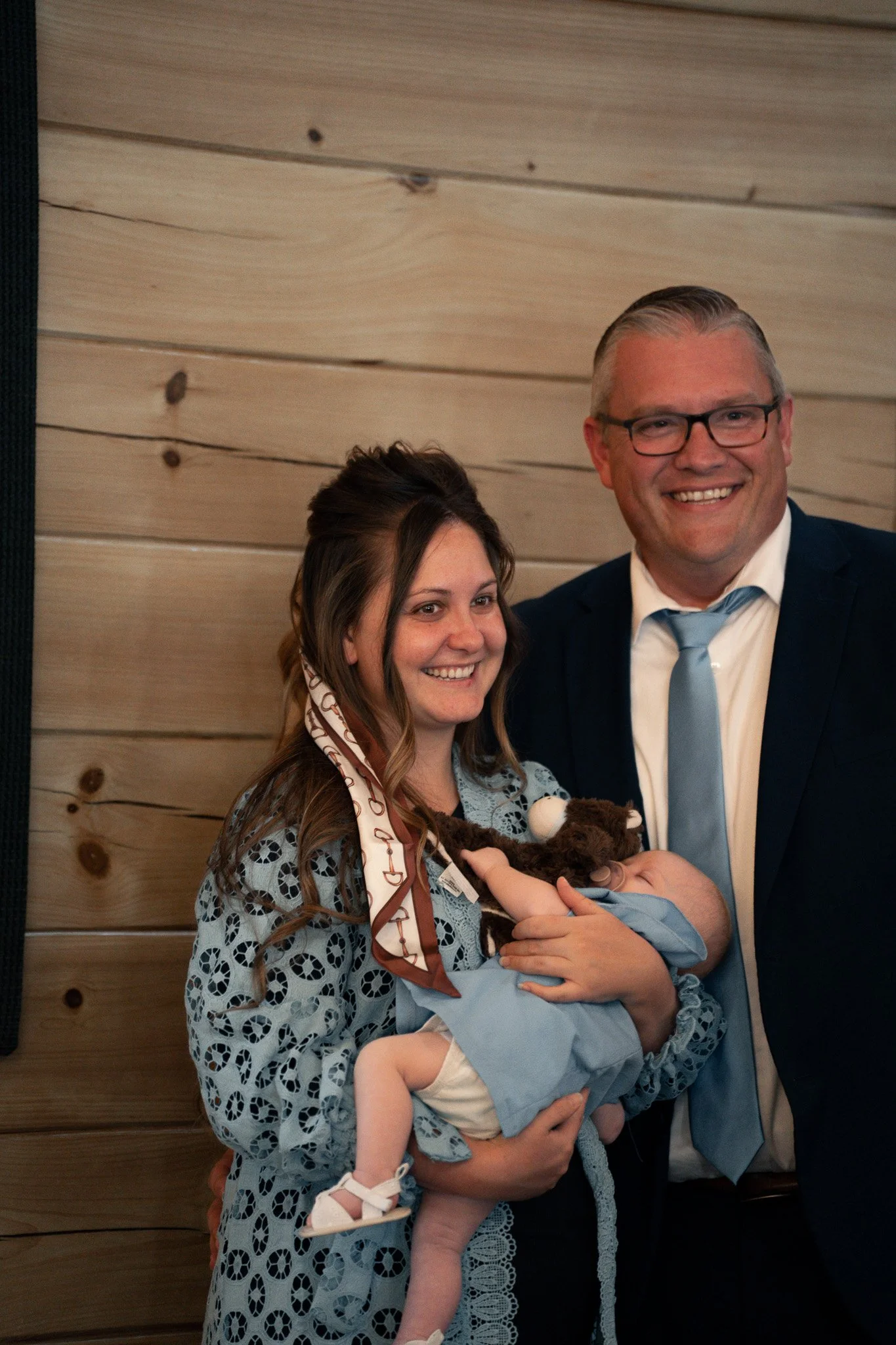 A woman holding a small child, both smiling, standing next to a man in a suit and tie, all posing in front of a wooden wall. The woman has long, wavy hair and wears a patterned dress, while the child is dressed in light blue and white clothing and ap