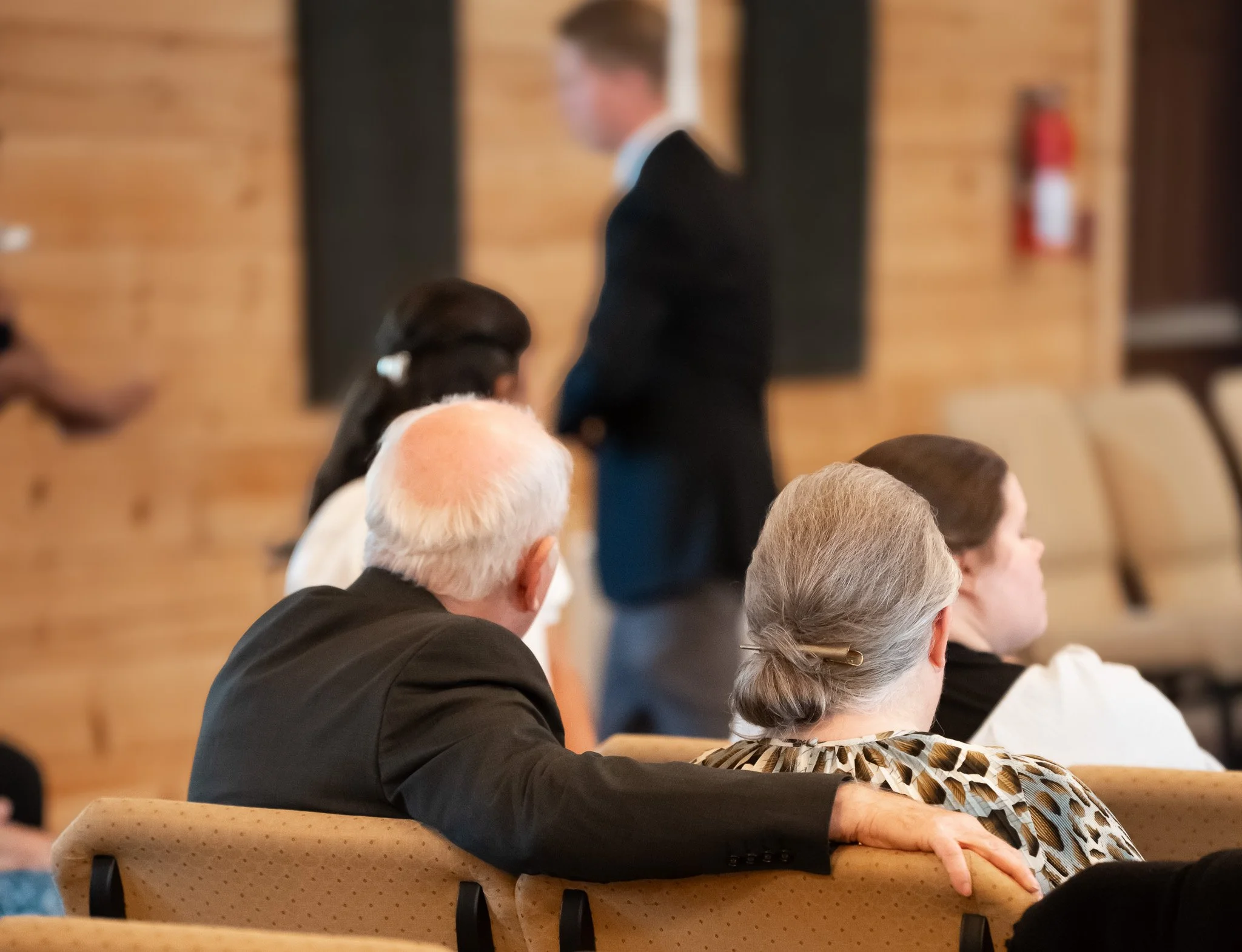 People seated at a wooden table attending an event, with some facing toward the front of the room. The background features a wooden wall, and a fire extinguisher is visible on the wall.