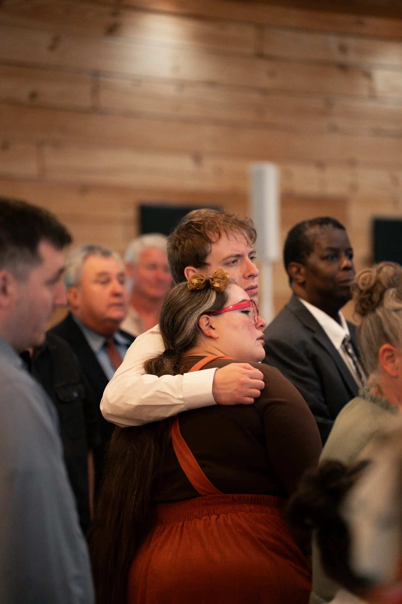 A group of people sitting closely together in an indoor setting, appearing to attend a formal event or gathering, with some individuals listening attentively.