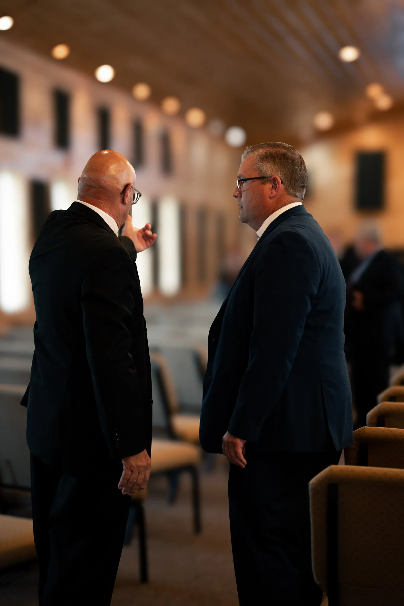 Two men in suits and glasses engaged in conversation inside a building with wooden ceiling and rows of chairs.