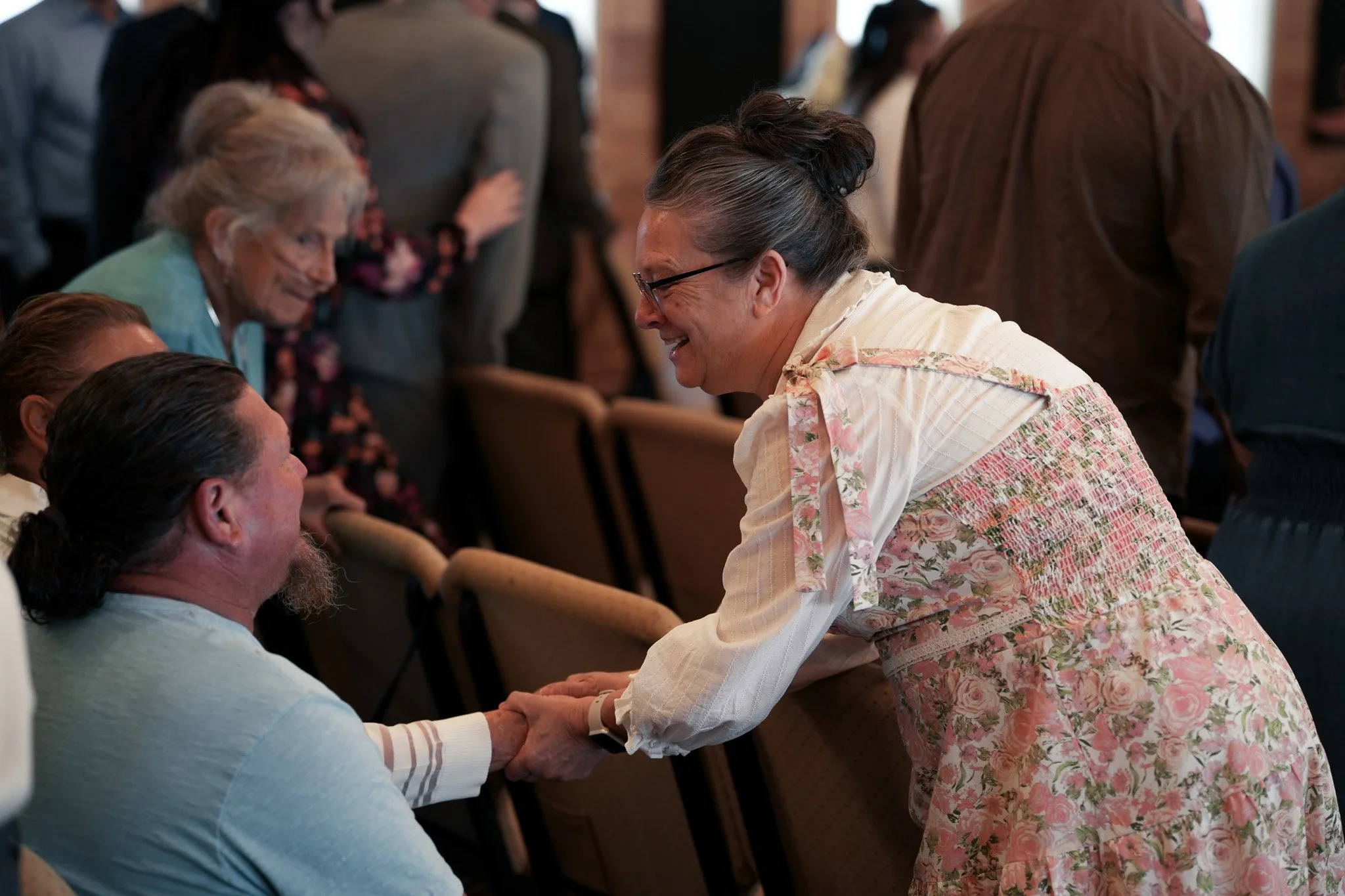 A woman in a floral dress and glasses is smiling and holding hands with a man with a beard, seated in a crowded indoor setting.