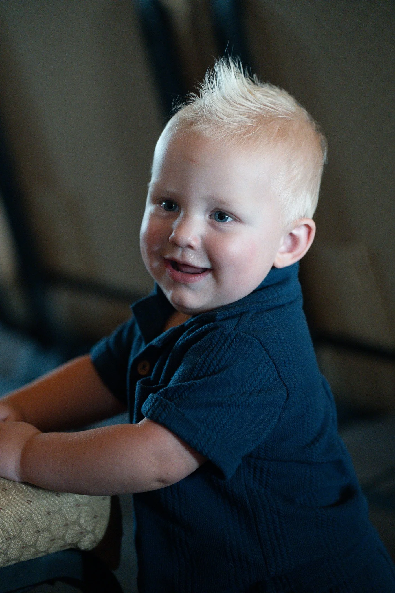 A young boy with blonde hair styled in a spiky manner, wearing a dark blue shirt, smiling and looking to the side.
