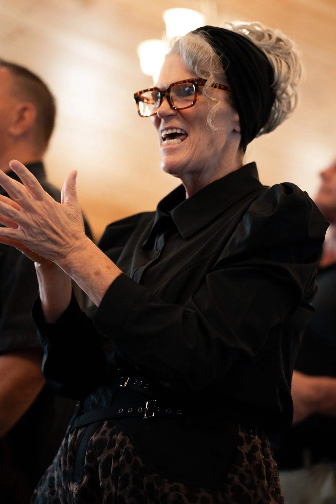 A woman with gray hair, wearing tortoiseshell glasses, a black headband, and a black top, laughing and gesturing with her hands at an indoor event.