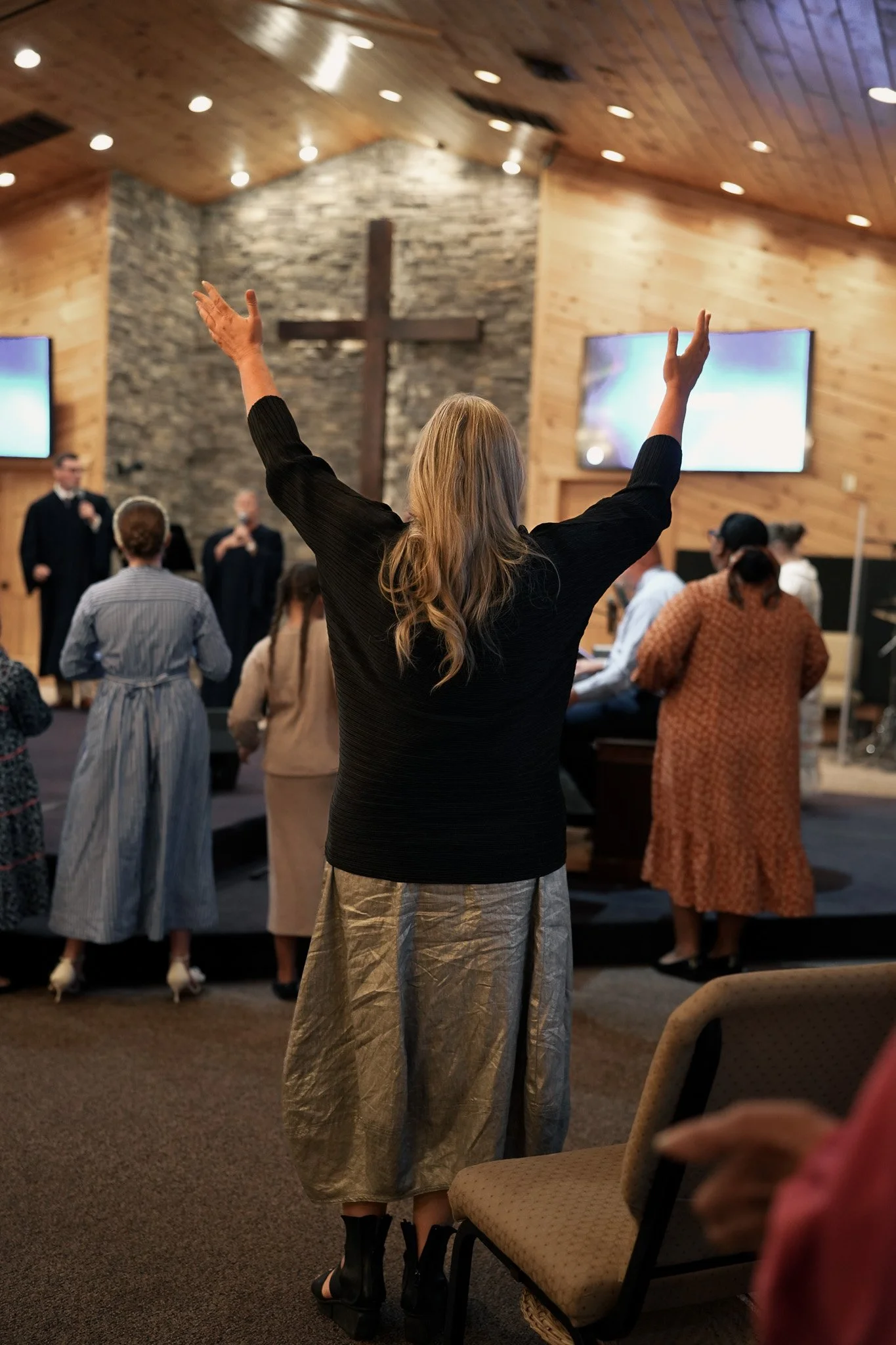A woman with long blond hair, wearing a black top and a shiny beige skirt, stands with arms raised in a church. The church interior has wooden walls, a cross on a stone wall background, and several people seated and standing during a service or praye