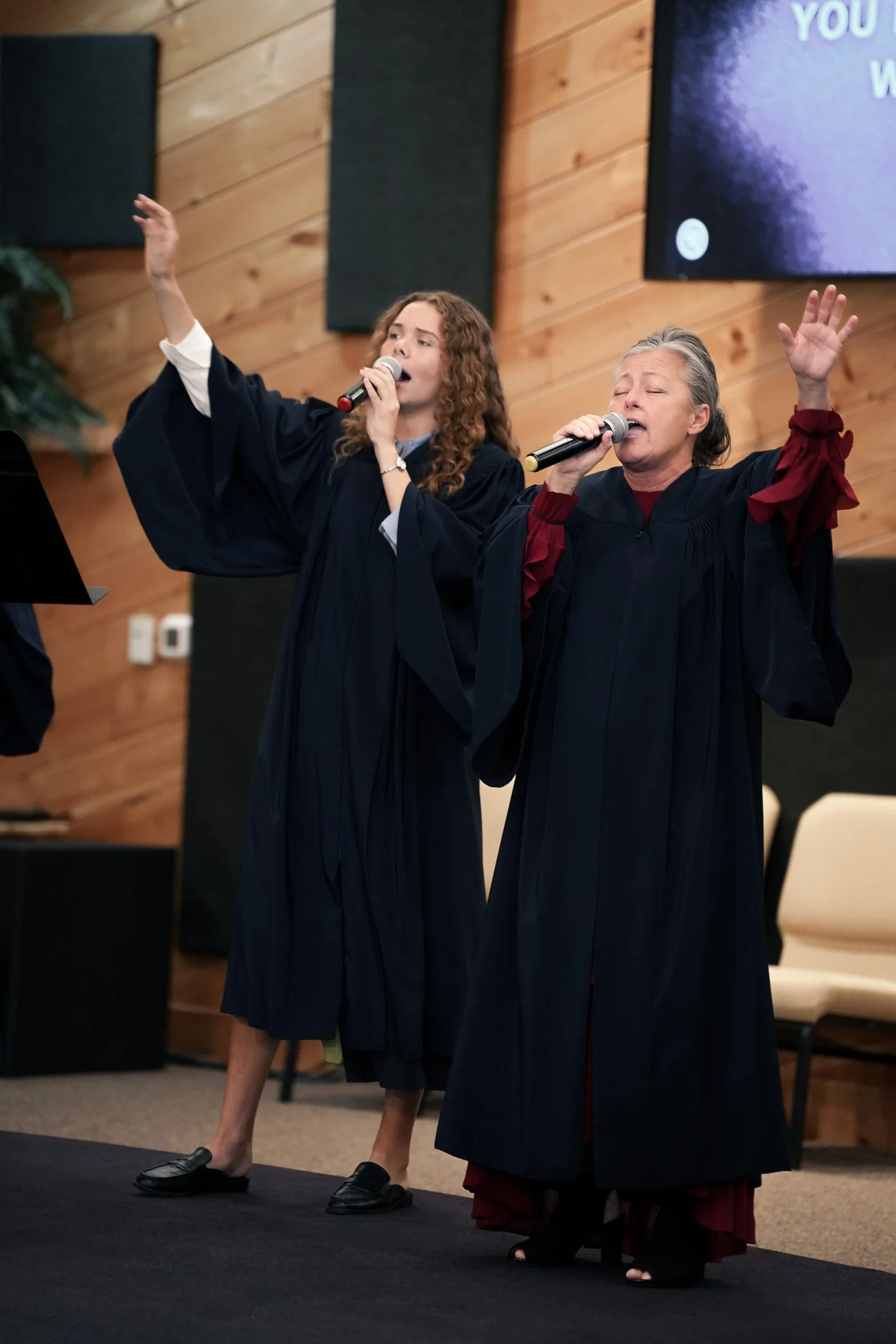 Two women dressed in choir robes with their eyes closed, singing and raising one hand each in a church or auditorium with wooden paneling on the walls.