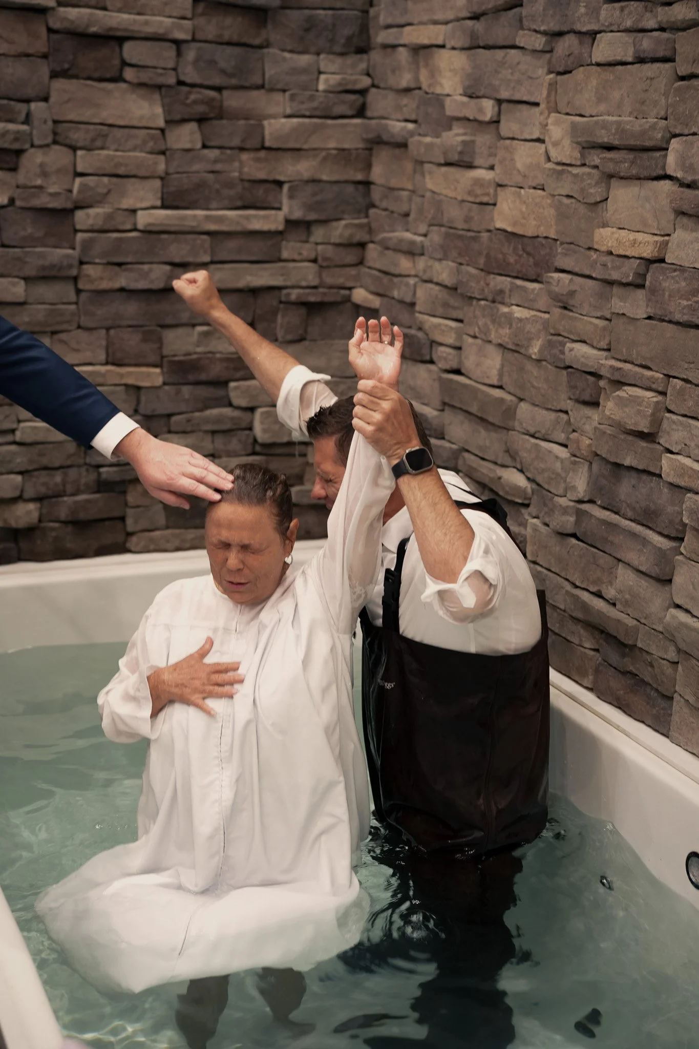 Two people are baptized in a bathtub filled with water, with one person raising their fist in the air and another person touching their head, while a third person's hand is on the person's head. The scene takes place indoors against a stone wall.