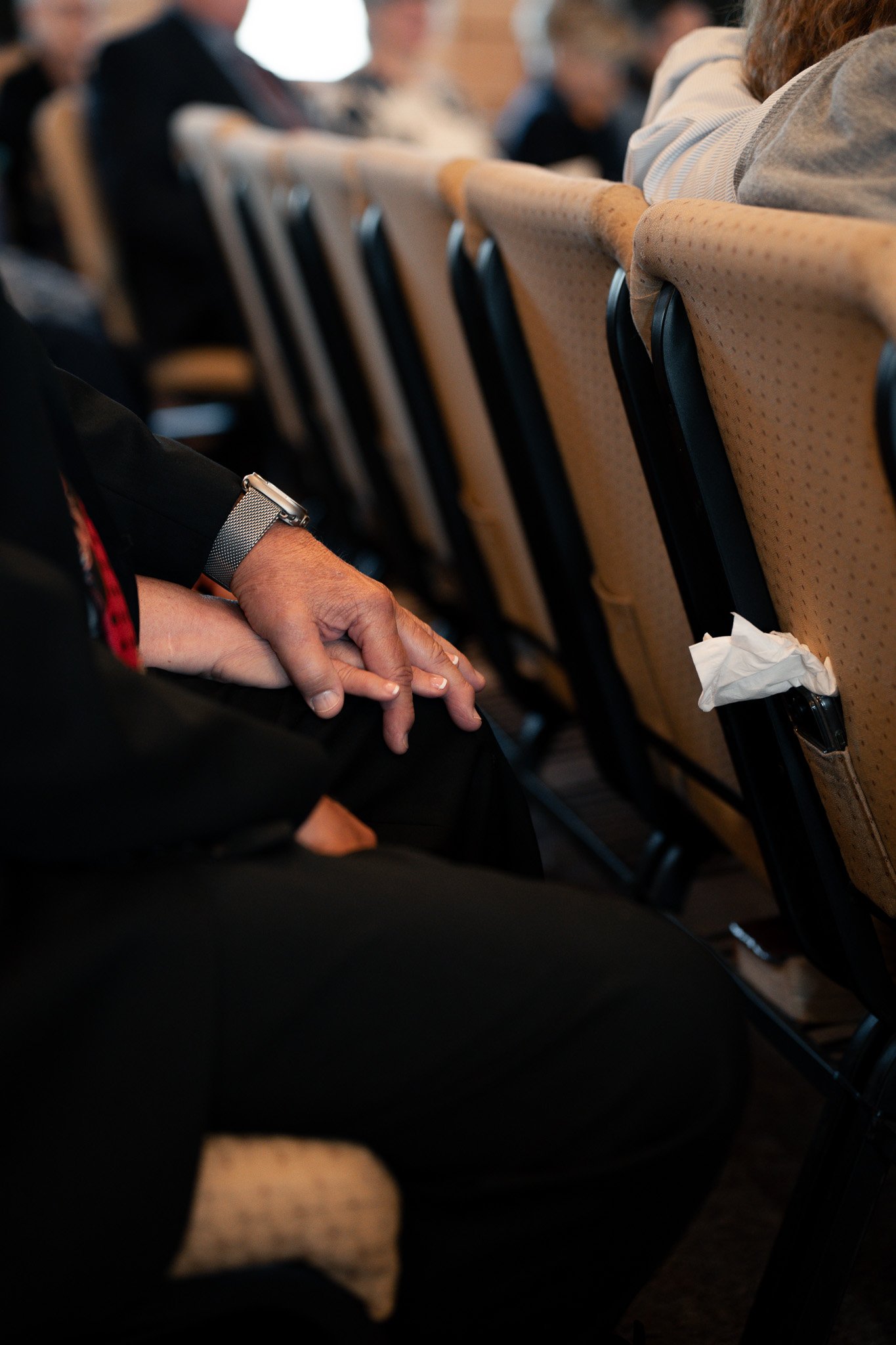 A person sitting in a conference room, holding their hands together on their lap, with a silver wristwatch on their left wrist. The background shows other people seated, with beige chairs and a tissue in the seat pocket.