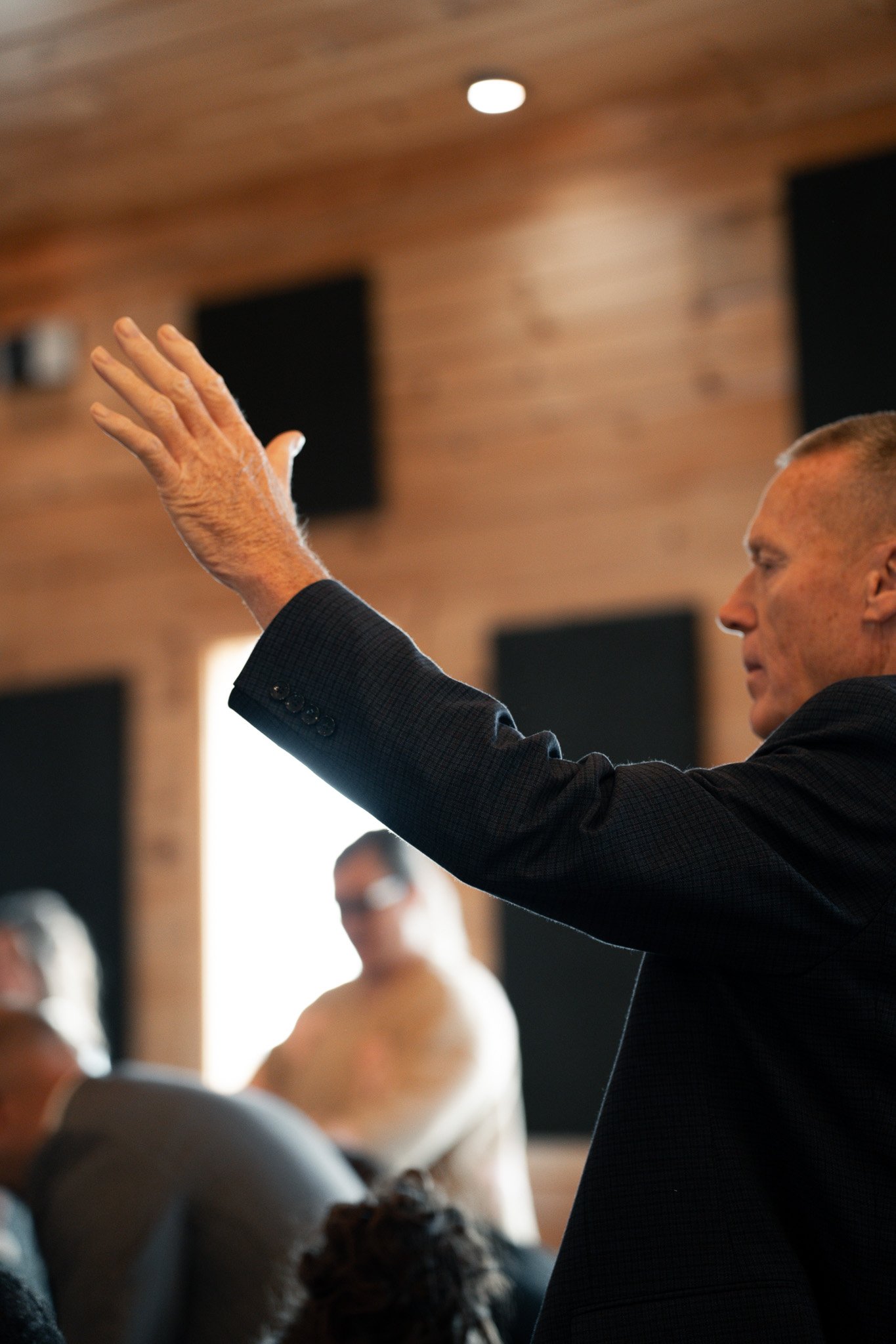A man in a black suit with short hair is raising his hand during a meeting or presentation in a room with wood panel walls. Several people are seated, some wearing masks.