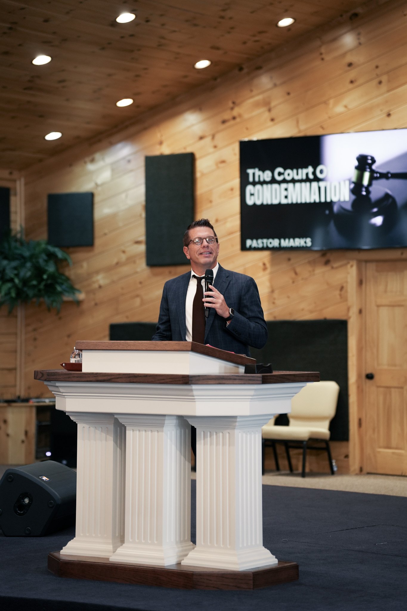 A man in a suit and glasses speaking into a microphone at a church or conference. Behind him is a black screen with white text that reads 'The Court of CONDEMNATION' and 'PASTOR MARK S'. The background features wooden paneling on the walls and ceilin