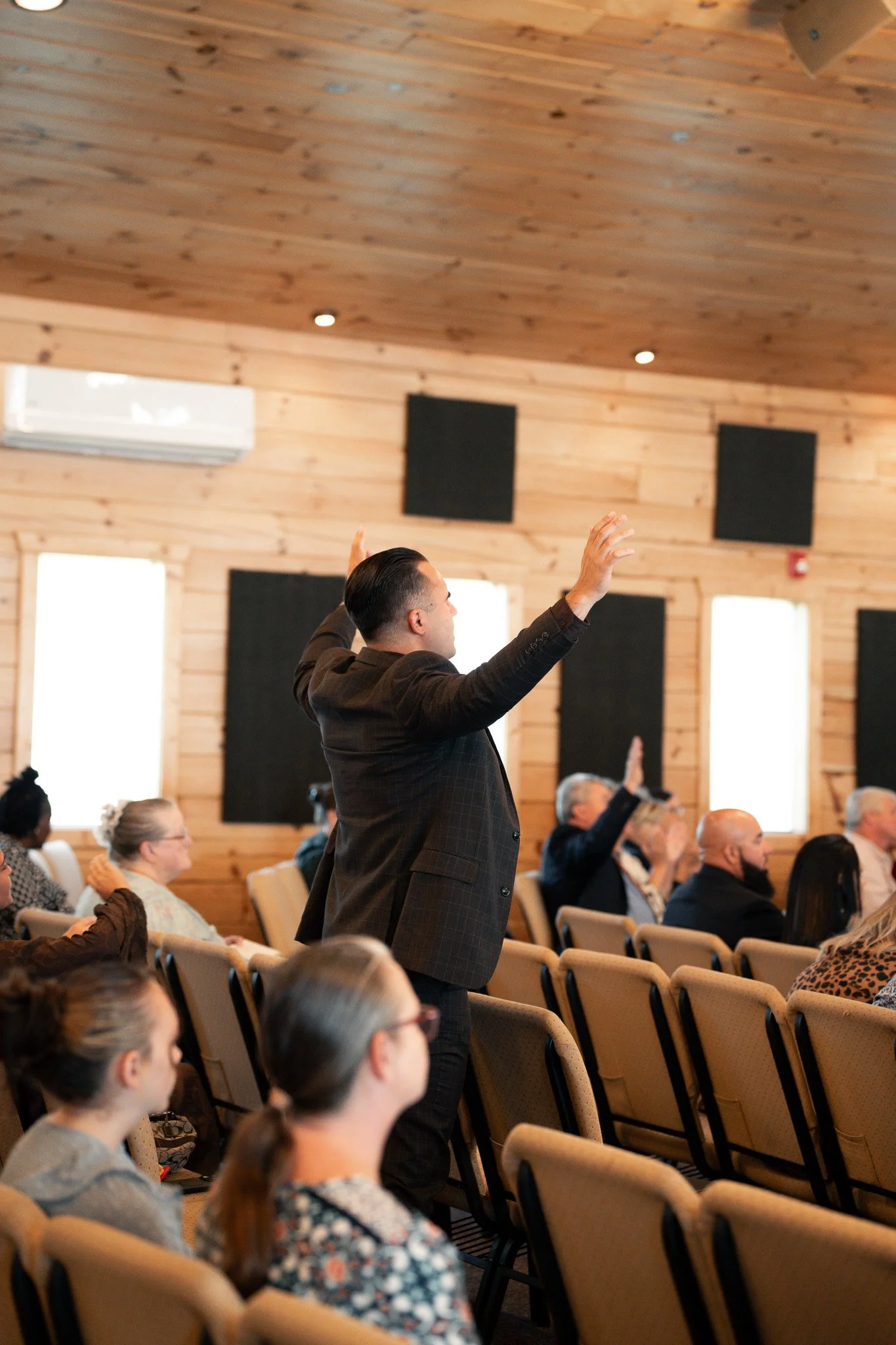 A man in a dark checkered suit is standing and raising his hands during a meeting or conference in a wood-paneled room. Several people are seated, some with hands raised, listening attentively.
