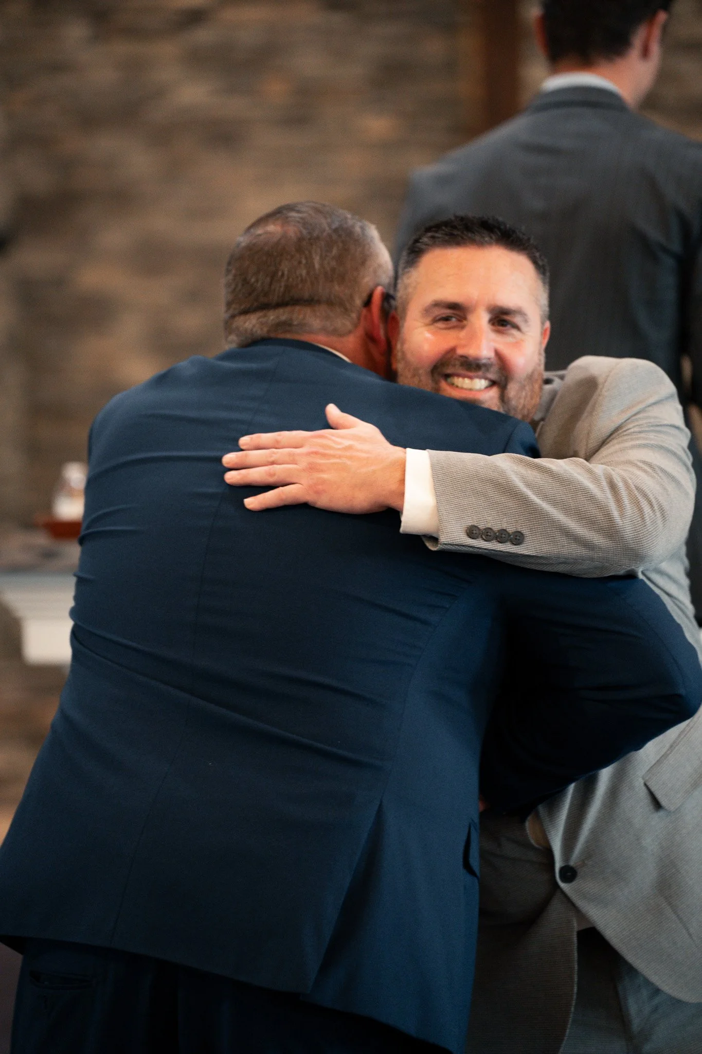 Two men dressed in suits hugging each other. The man facing the camera has a beard and is smiling. The other man has his back to the camera and appears to be older with gray hair.