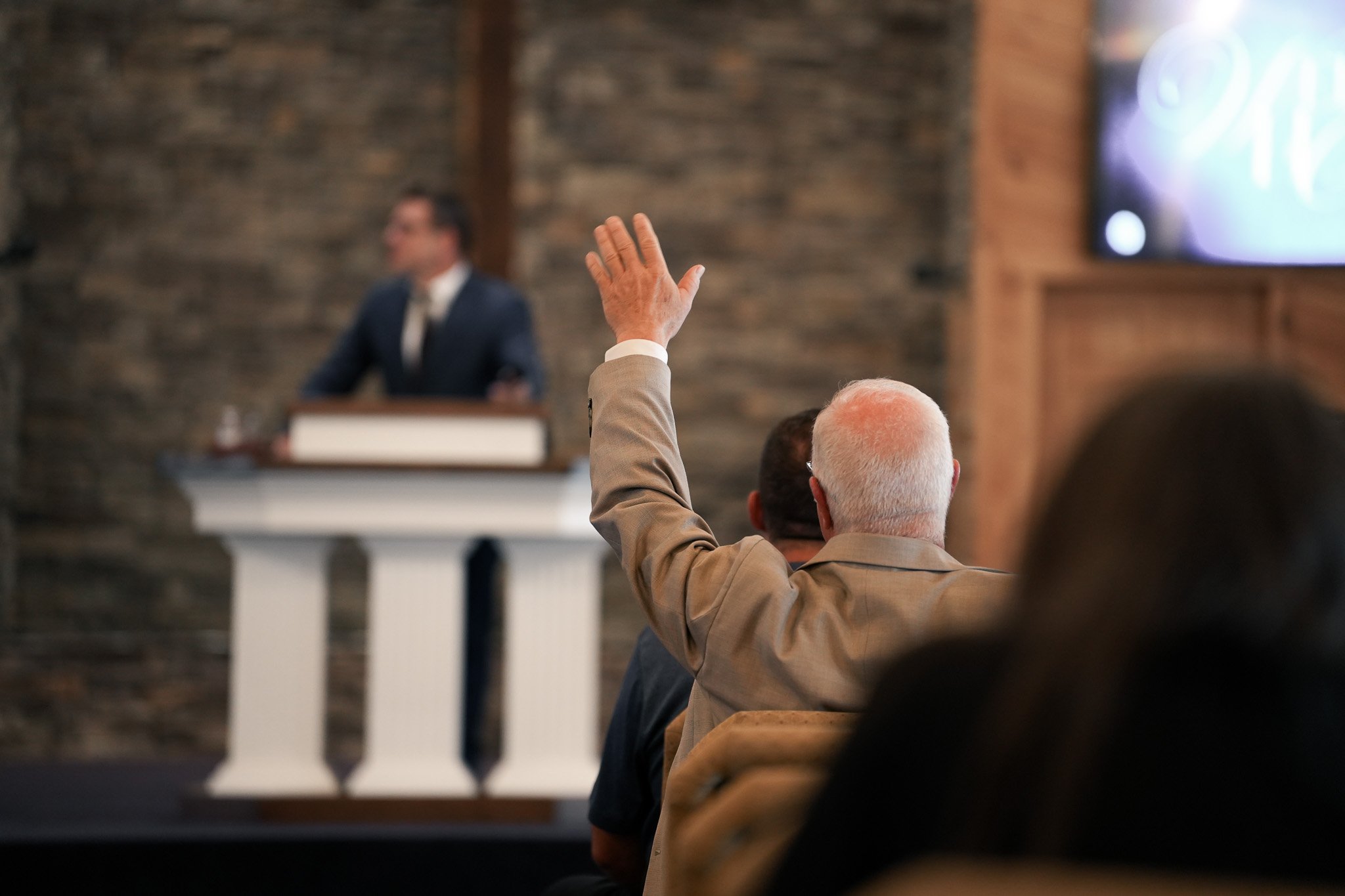 A person with a raised hand in a beige blazer in a room with a stone wall and a speaker at a podium, blurred man in a suit is in the background, and an audience member is visible in the foreground.