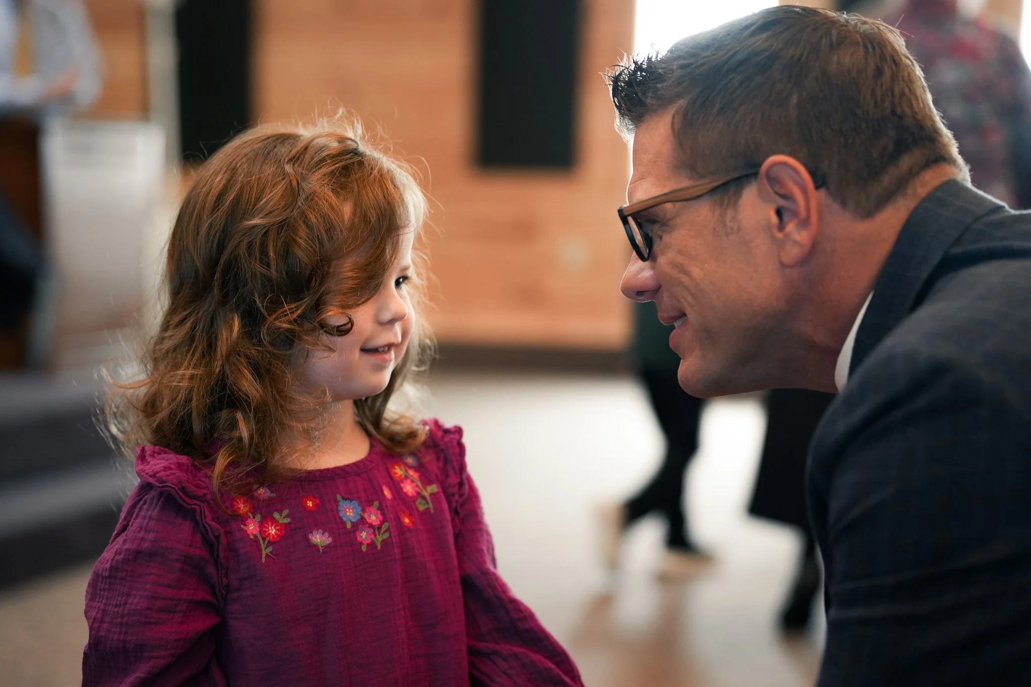 A young girl with red hair and a pink embroidered dress is smiling and looking at a man with glasses, who is smiling back at her.