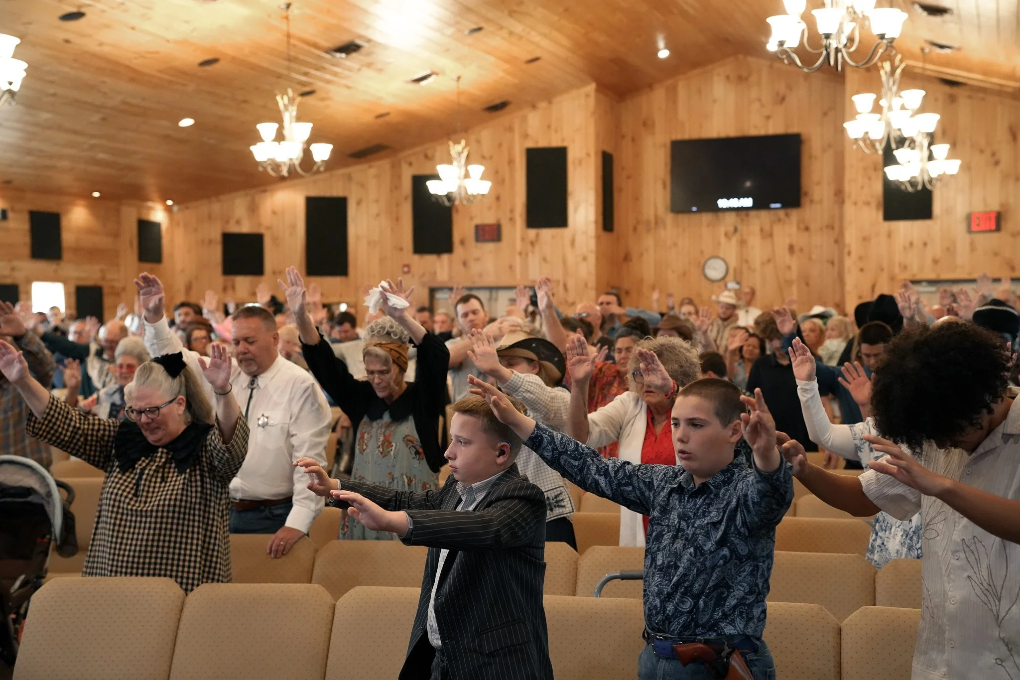 A large group of people in a wooden church or community hall raising their hands during a prayer or community event, with chandeliers hanging from the ceiling and a television screen on the wall.