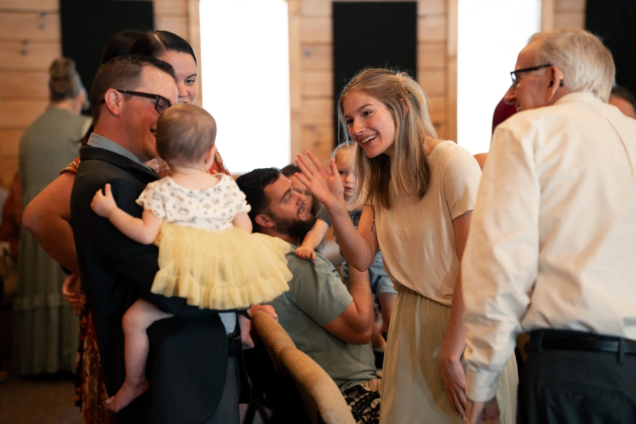 A group of people, including children and adults, are gathered in a circle, engaging in conversation and social interaction in an indoor setting with wooden walls. The scene shows a woman smiling and gesturing as she talks to an elderly man and other