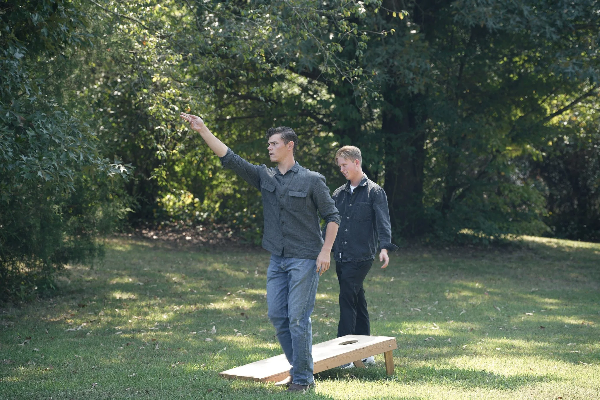 Two young men play cornhole in a park with green trees in the background. One man is throwing a bean bag while the other watches.