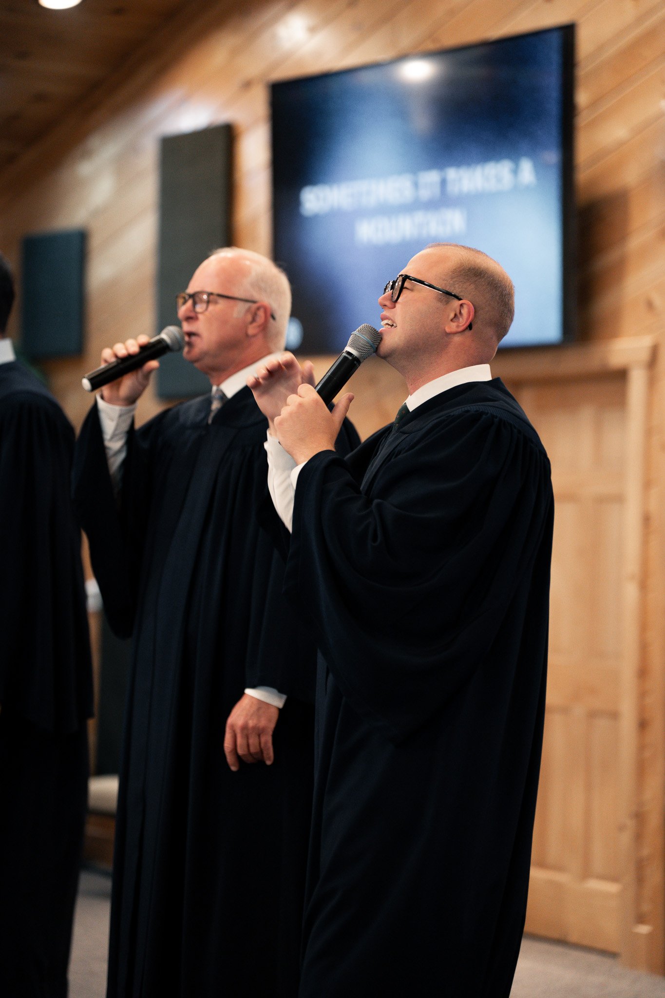 Two men in suits and glasses holding microphones, participating in a formal event in a wooden room. One man has a gray bald head, and the other is younger with a shaved head. A large screen behind them displays text that is partially visible.