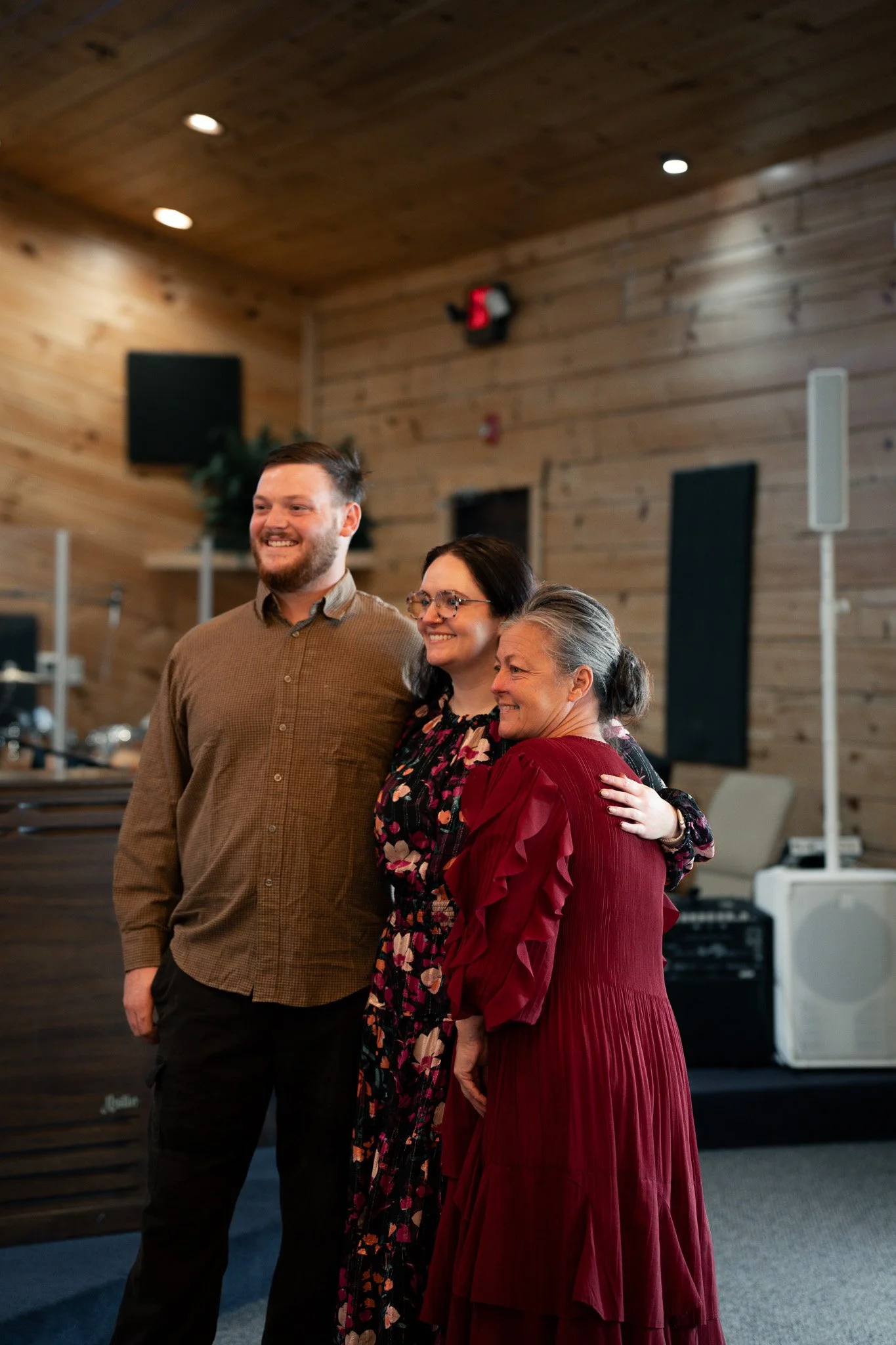 Three people standing in a room with wooden walls, smiling and posing for a photo. Two women and one man, with a music speaker and equipment visible in the background.