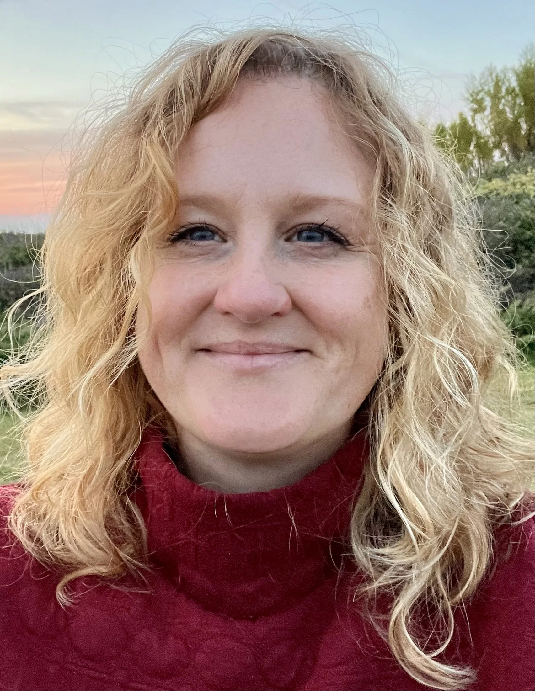 Close-up of Melissa's face, with blonde curly hair, smiling and wearing a red sweater, outdoors during sunset with trees in the background.