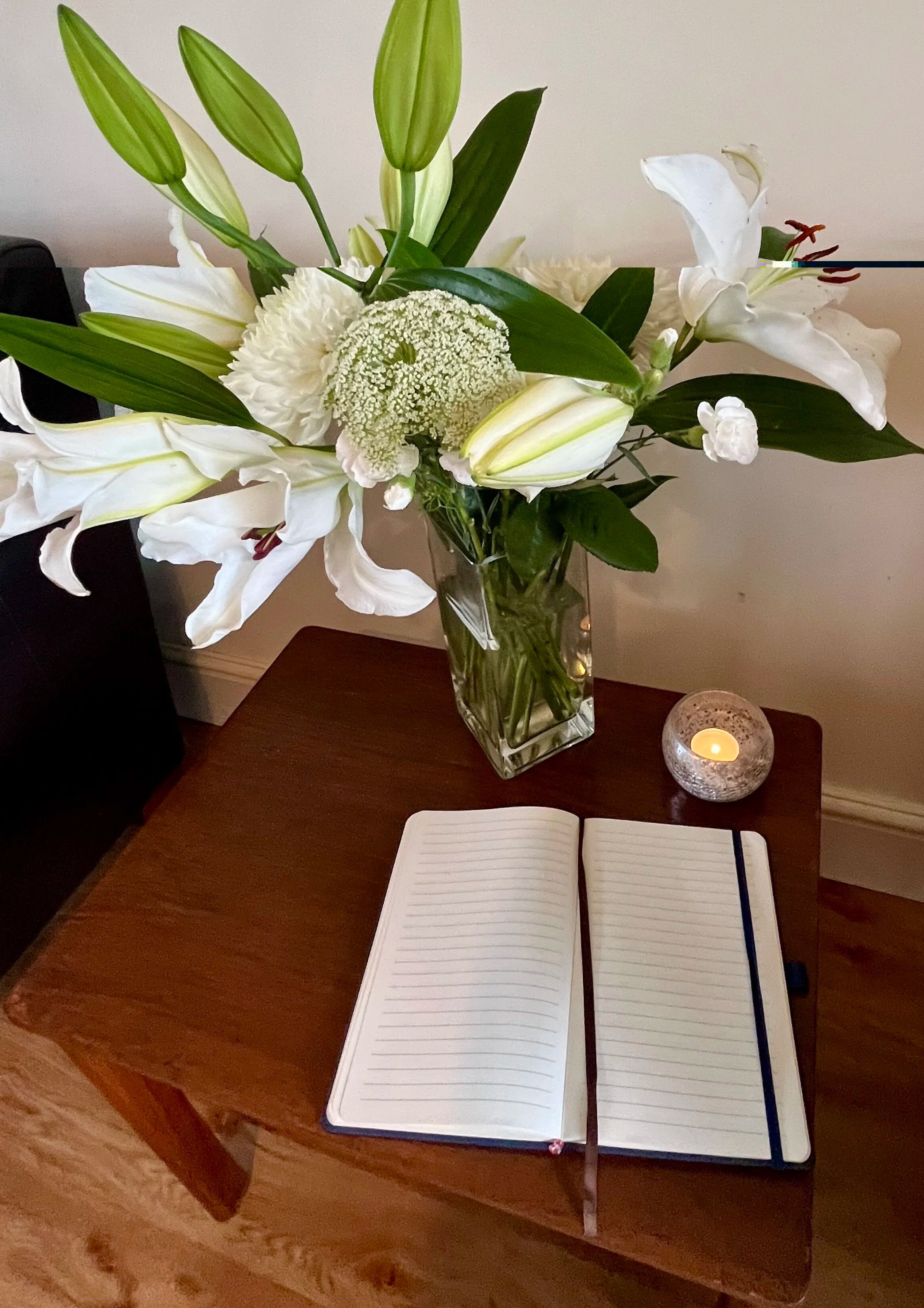 A wooden table with an open lined notebook, a glass vase with white flowers and green leaves, a small candle holder, and a partially visible black electronic device.