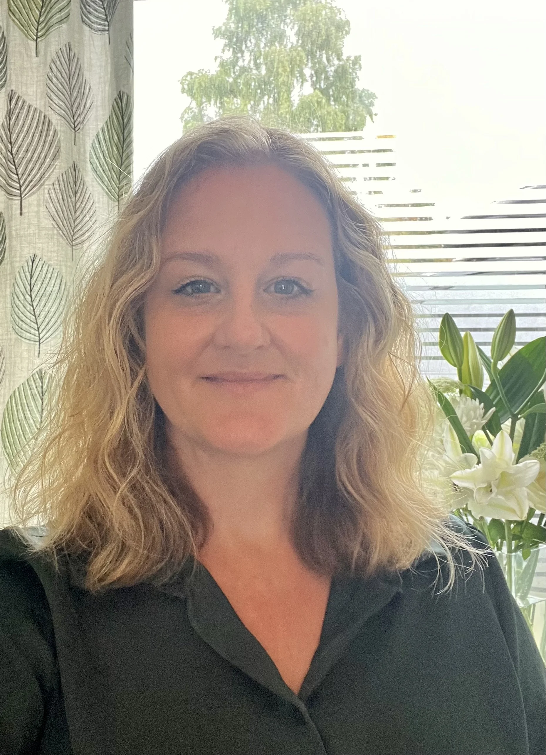 Melissa with curly blonde hair and blue eyes sitting indoors near a window, with green leaves and white flowers in the background.