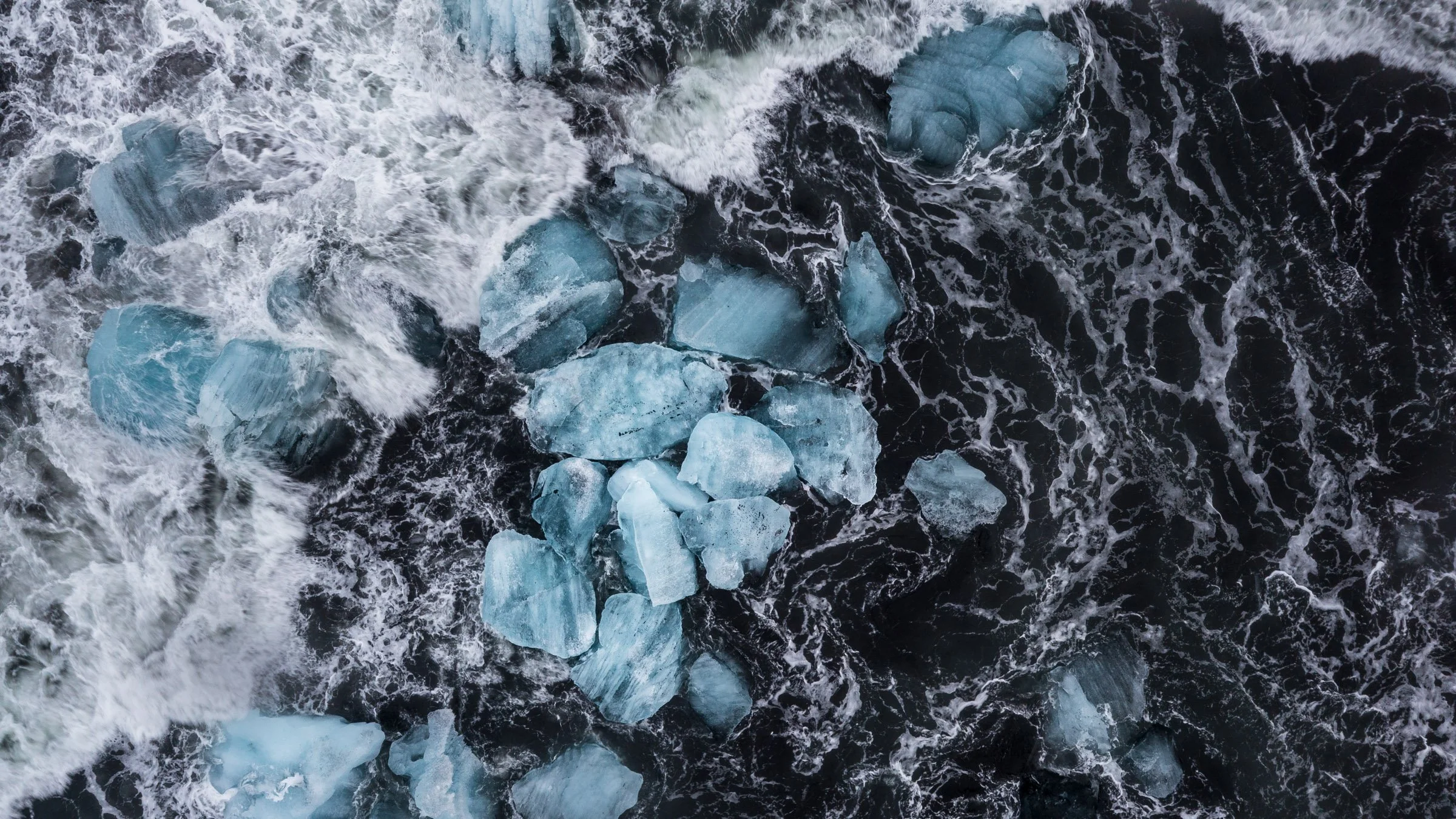 Aerial view of icy blue icebergs surrounded by dark ocean water with waves crashing around.