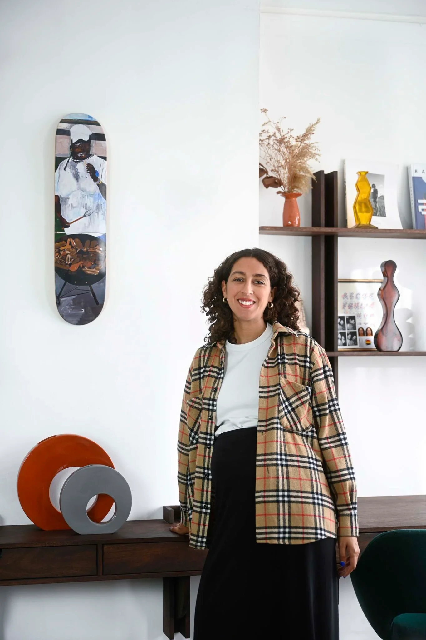 Une femme souriante avec des cheveux bouclés, portant un haut blanc et un pantalon noir, chemise à carreaux beige et rouge et un sac noir, debout dans une pièce décorée avec des œuvres d'art modernes, une étagère avec des livres, des vases et des cadres photos.
