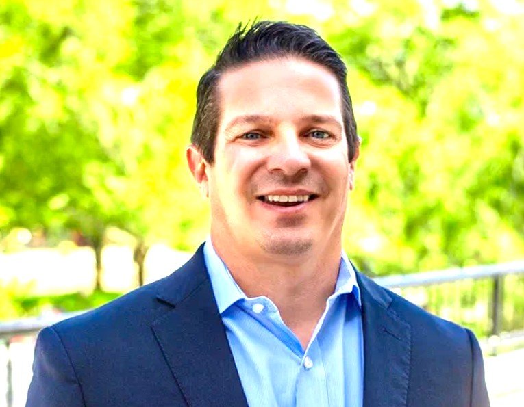 A smiling man in a dark blue suit and light blue shirt standing outdoors with green trees and a metal railing in the background.