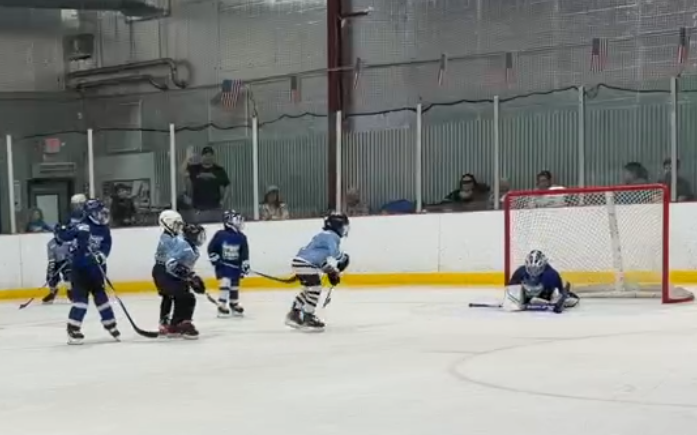 Youth hockey game with kids in blue jerseys playing on an ice rink, goalie in position near the net, and spectators watching behind the glass.