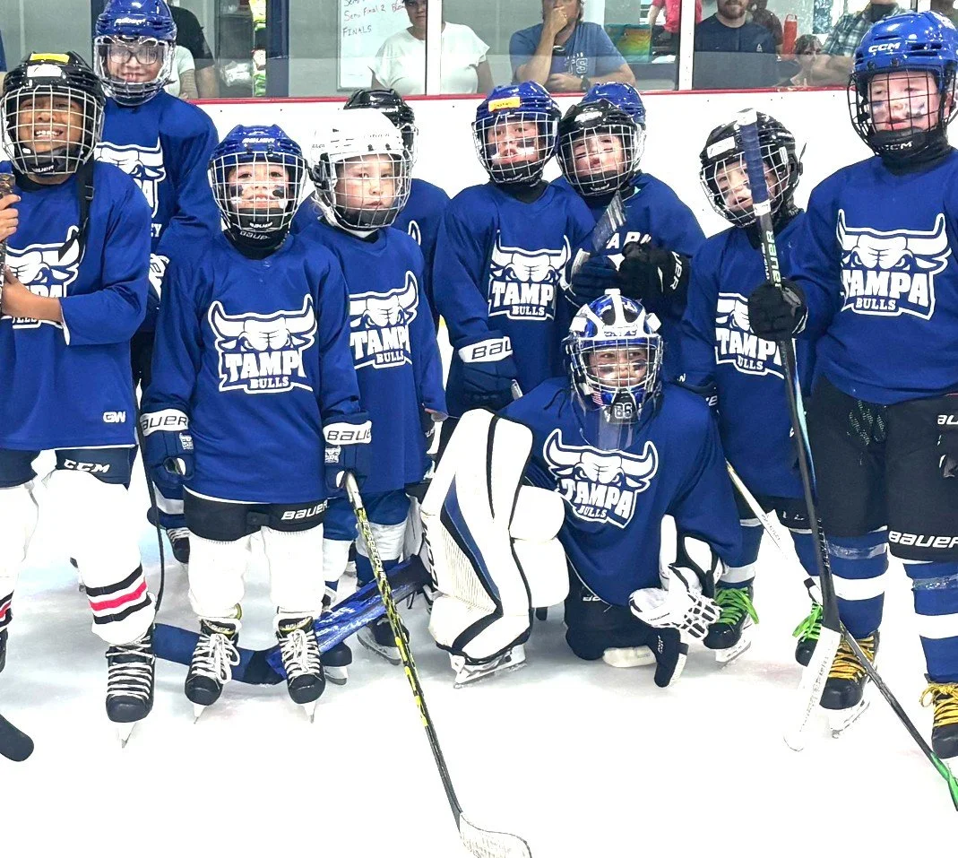 Group of kids in blue hockey jerseys with 'TAMPA BULLS' logo, wearing helmets and hockey gear, posing on ice rink.