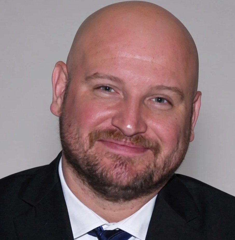 A bald man with a beard and mustache, wearing a black suit and a white dress shirt, smiling at the camera.