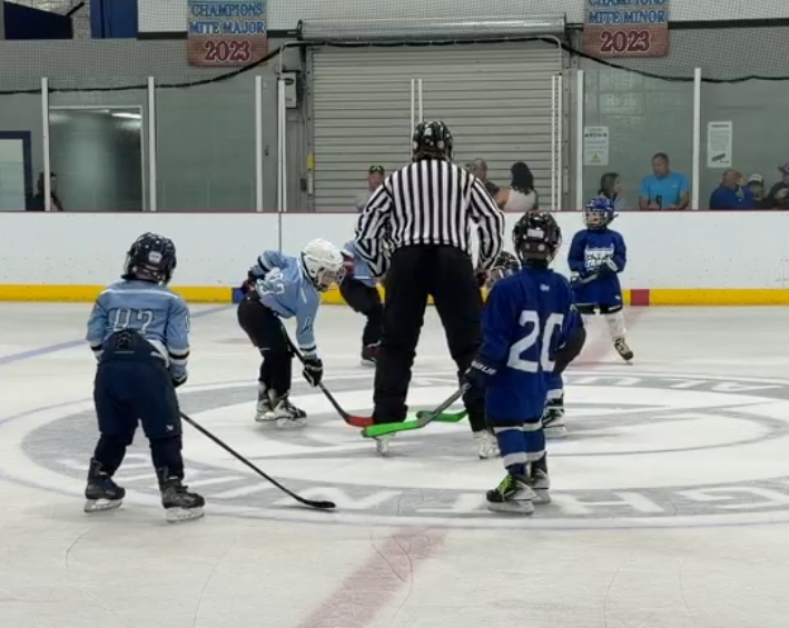 Kids playing hockey on ice rink with referee at center, preparing for faceoff, with spectators watching behind glass.