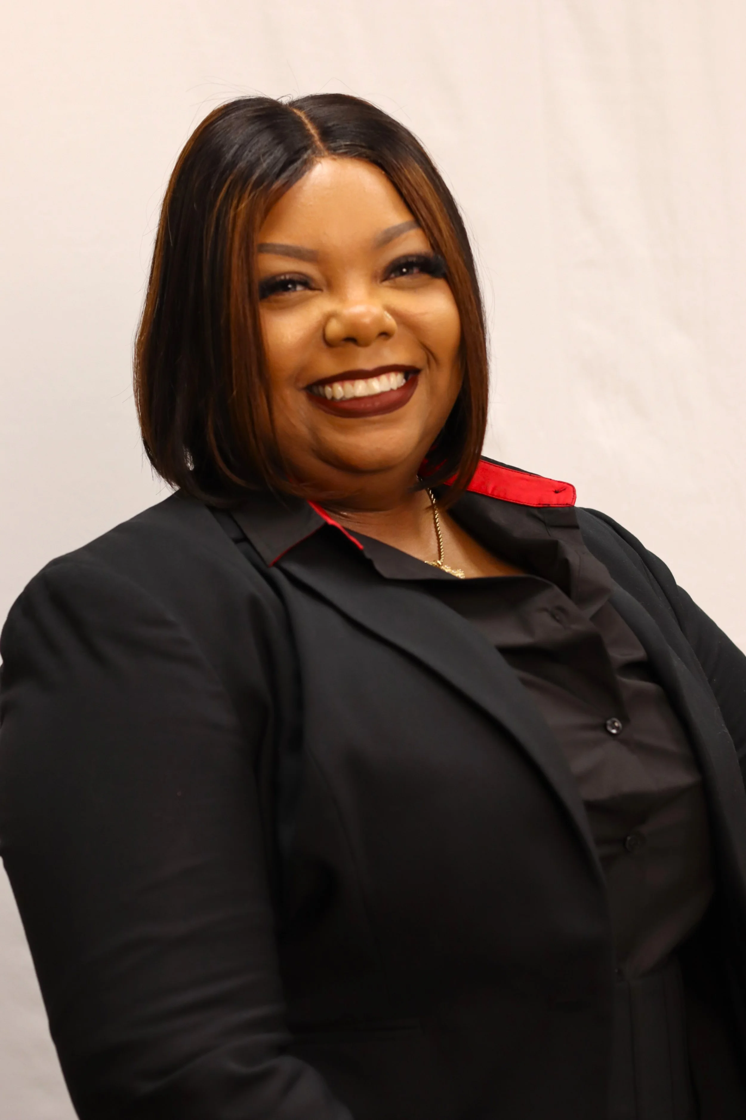 A woman with medium-length brown hair and dark lipstick smiling, wearing a black jacket with red accents on the collar, standing against a plain light-colored background.
