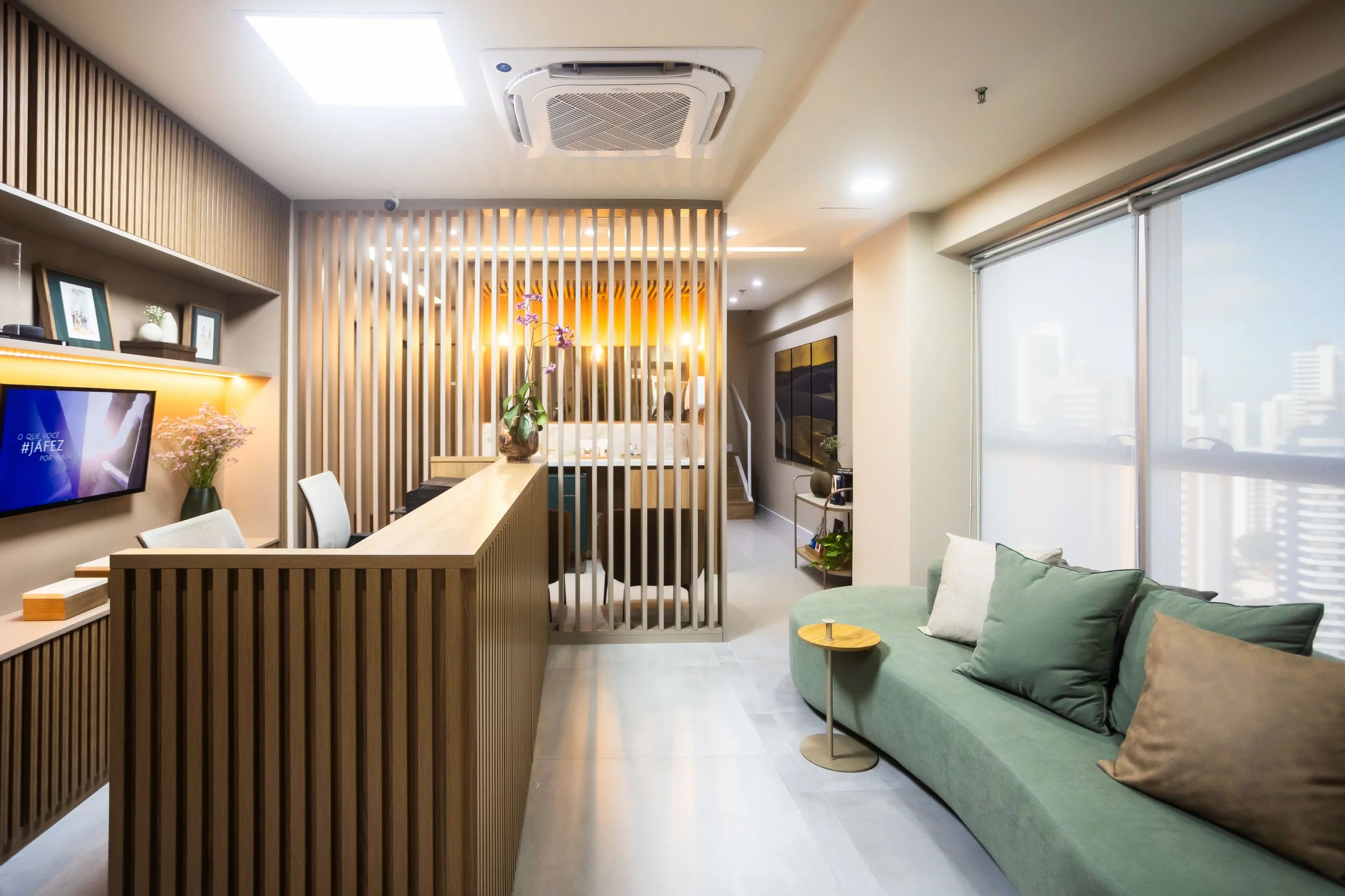Sala moderna com sofá verde, poltrona, televisão de tela plana na parede, mesa com plantas e decoração, divisória de madeira, iluminação suave e grandes janelas de vidro com cortinas parcialmente fechadas.