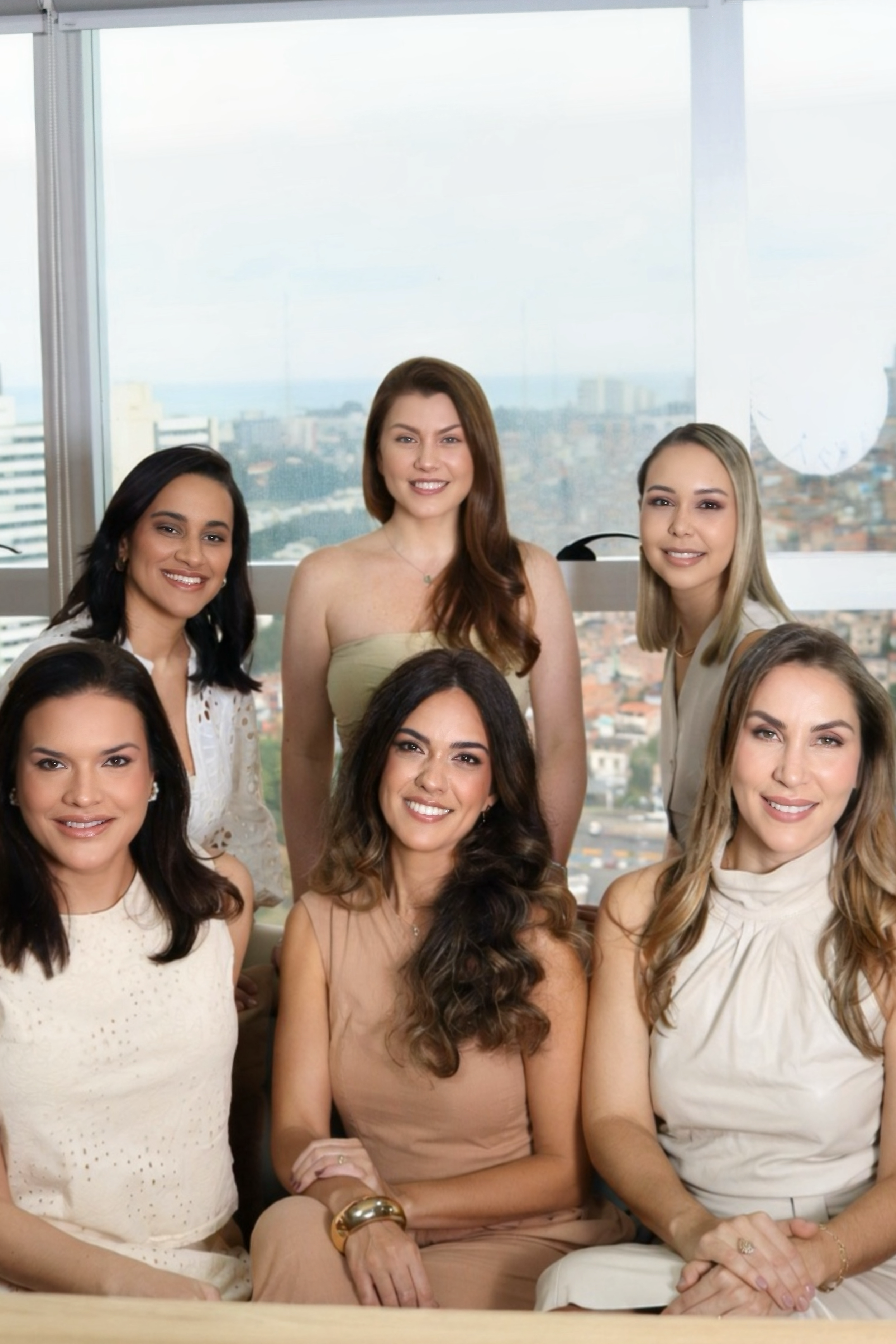 Mulheres sorrindo e posando juntas em um ambiente de escritório com vista para a cidade ao fundo.