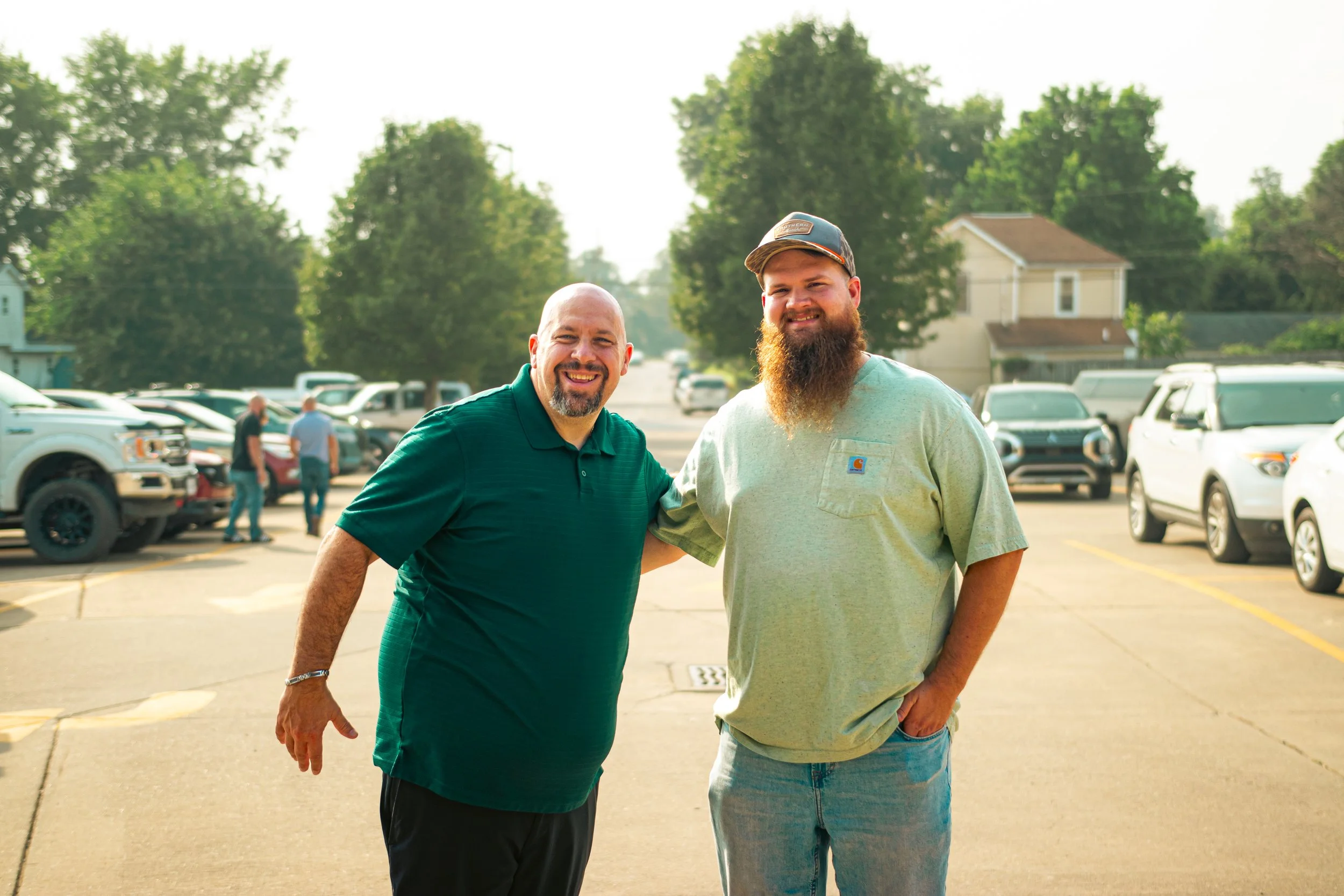 Two smiling men with arms around each other standing in a parking lot with trees and houses in the background, on a sunny day.